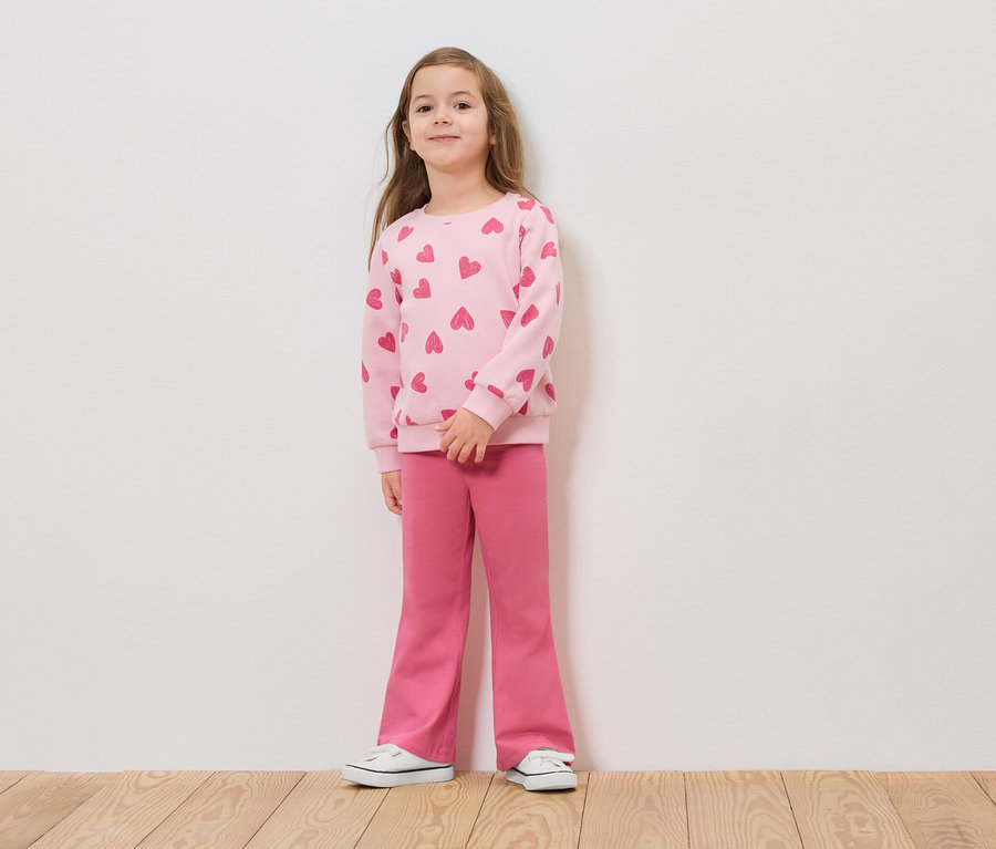 Dziewczynka stoi przed białą ścianą w różowym swetrze w serca i różowych spodniach.