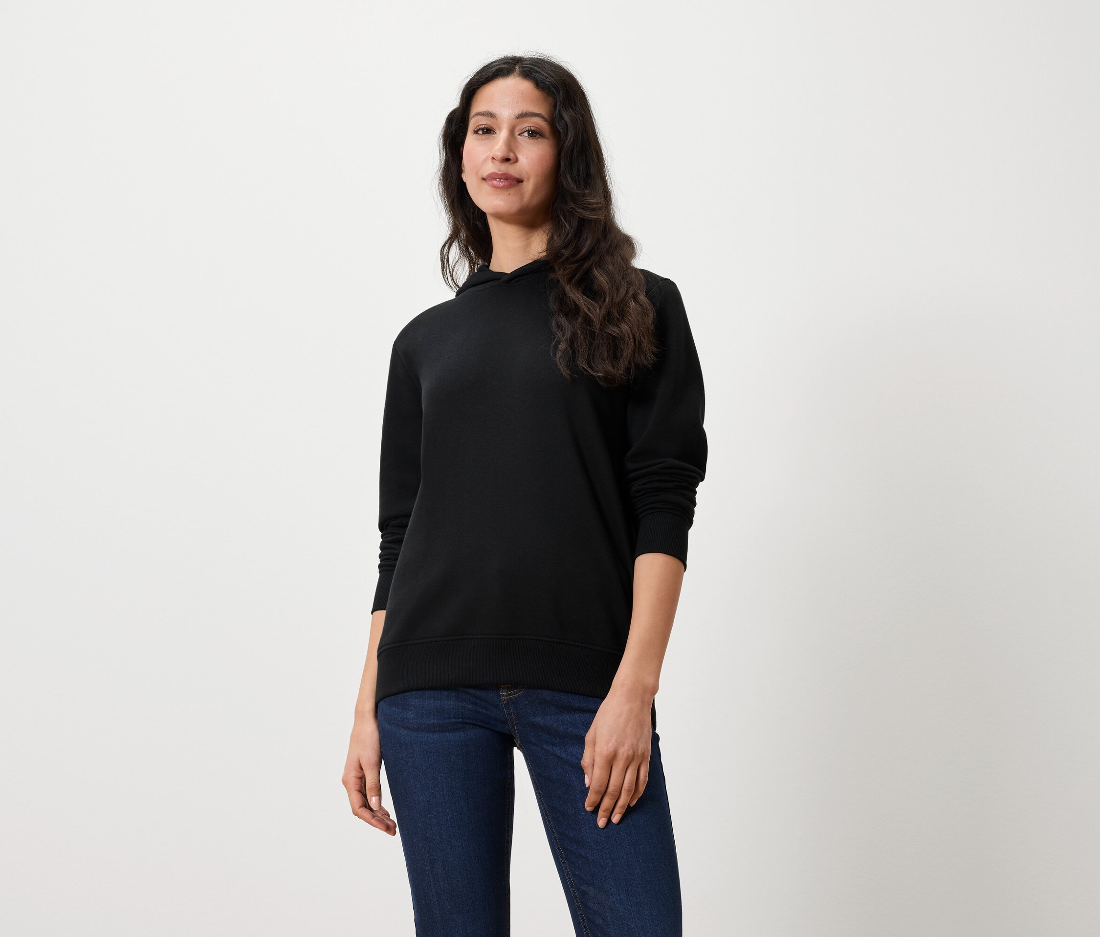 Kobieta z długimi brązowymi włosami stoi i ma na sobie czarną bluzę z kapturem i dżinsy.