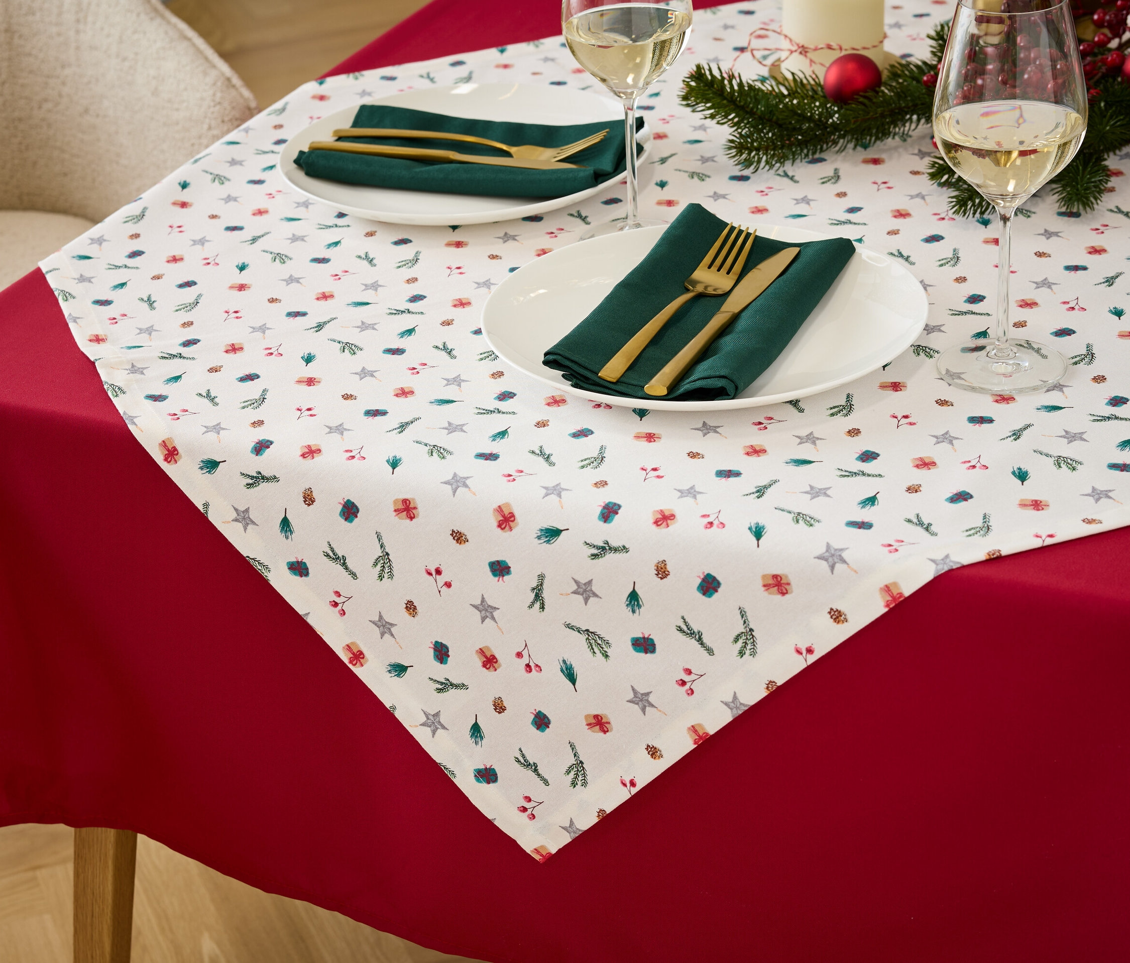 Detal stolu z czerwonym obrusem, białym bieżnikiem ze świątecznym motywem, zielonymi serwetkami z materiału i złotymi sztućcami.