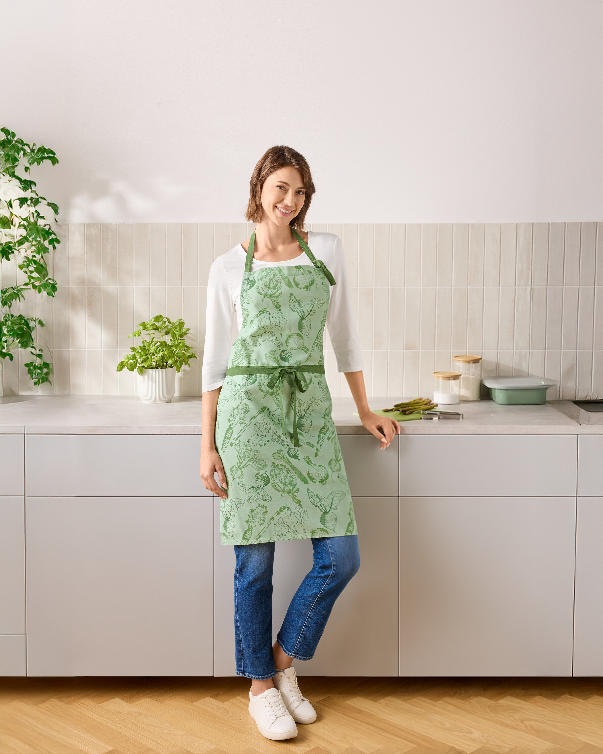 Kobieta stoi w kuchni, ubrana w zielony fartuch z motywem warzyw.