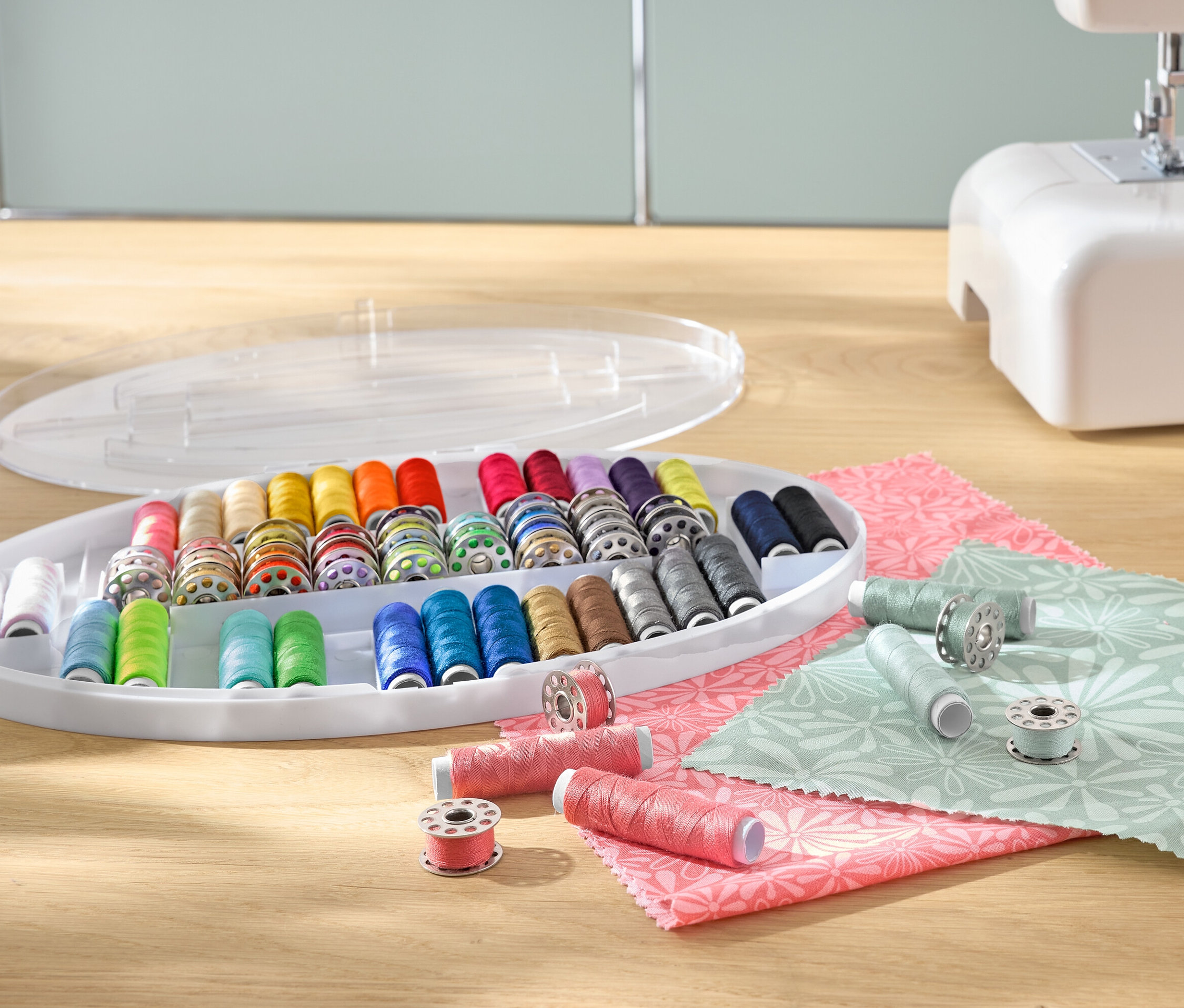 Zestaw nici do szycia z kolorowymi szpulami nici w plastikowym organizerze, obok maszyny do szycia i materiałów.