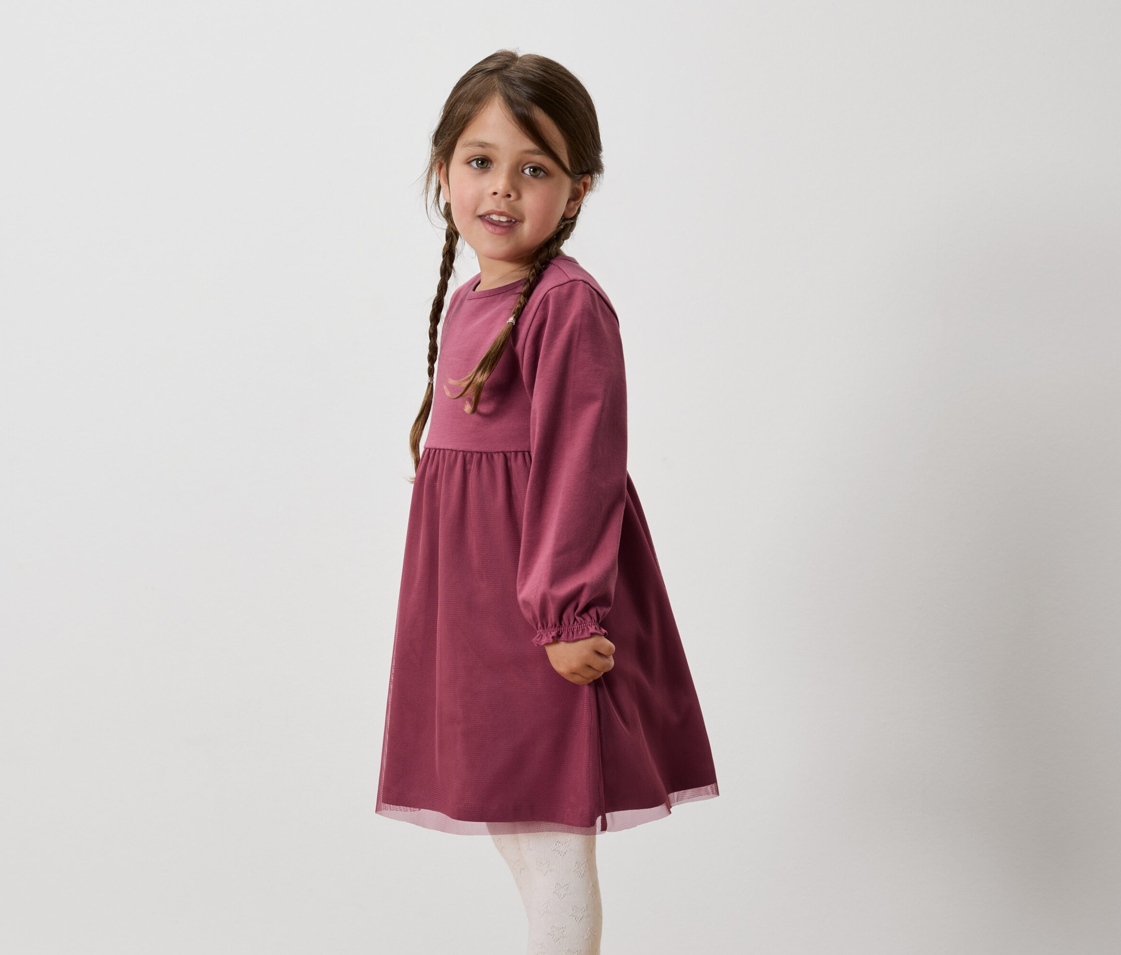Dziewczynka z brązowymi włosami splecionymi w warkocze ma na sobie bordową sukienkę z długim rękawem i białe legginsy.