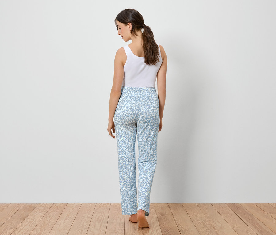 Kobieta od tyłu, ubrana w biały podkoszulek i jasnoniebieskie spodnie w kwiatowy wzór, stoi na drewnianej podłodze.