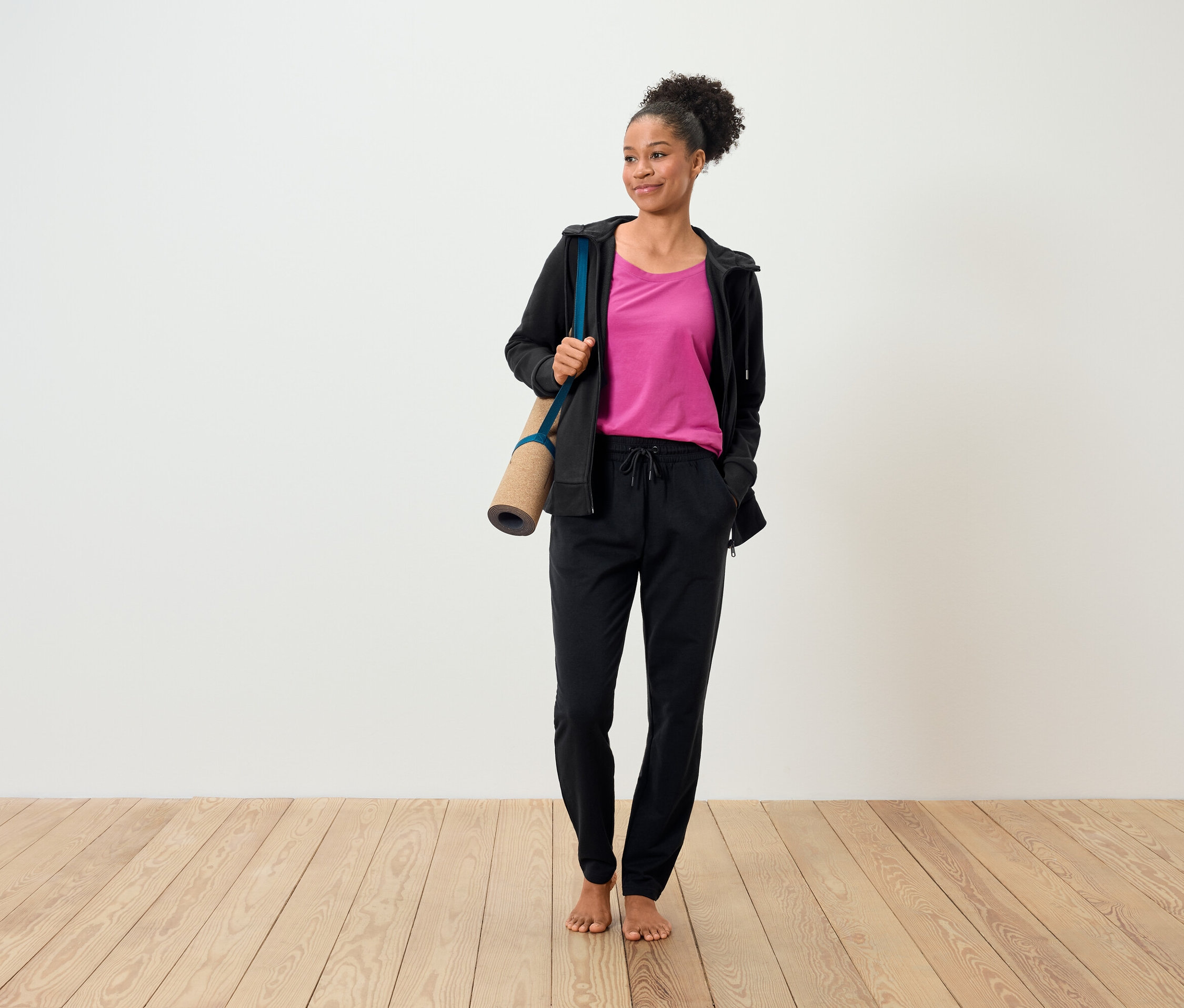 Kobieta z brązowymi włosami trzyma matę do jogi i ma na sobie różowy t-shirt, czarną bluzę i czarne spodnie.