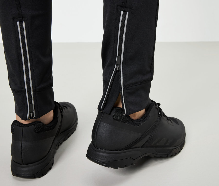 Nogi w czarnych butach sportowych i czarnych spodniach ze srebrnymi zamkami błyskawicznymi na szarym tle.