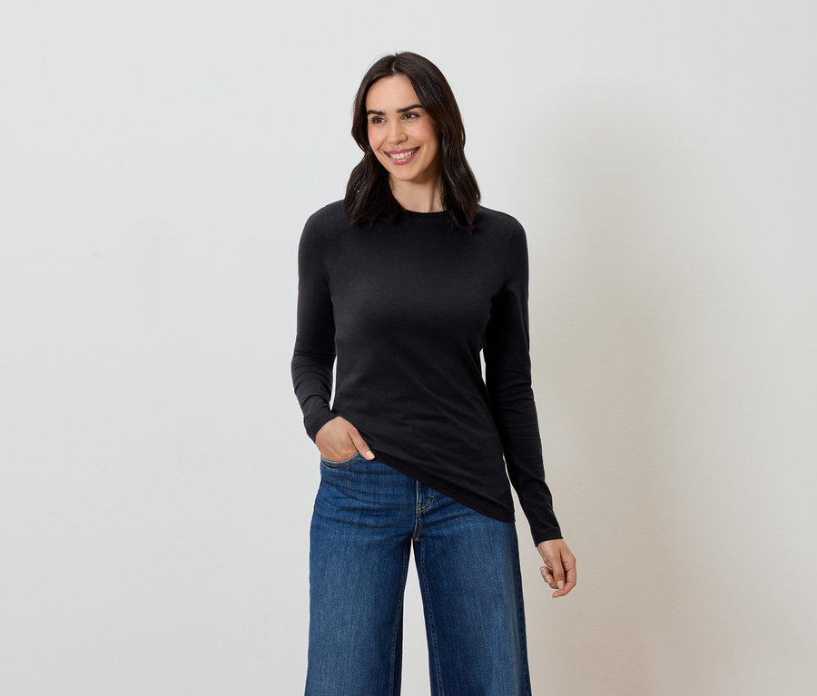 Kobieta z brązowymi włosami pozuje w czarnej bluzce z długim rękawem i niebieskich dżinsach.