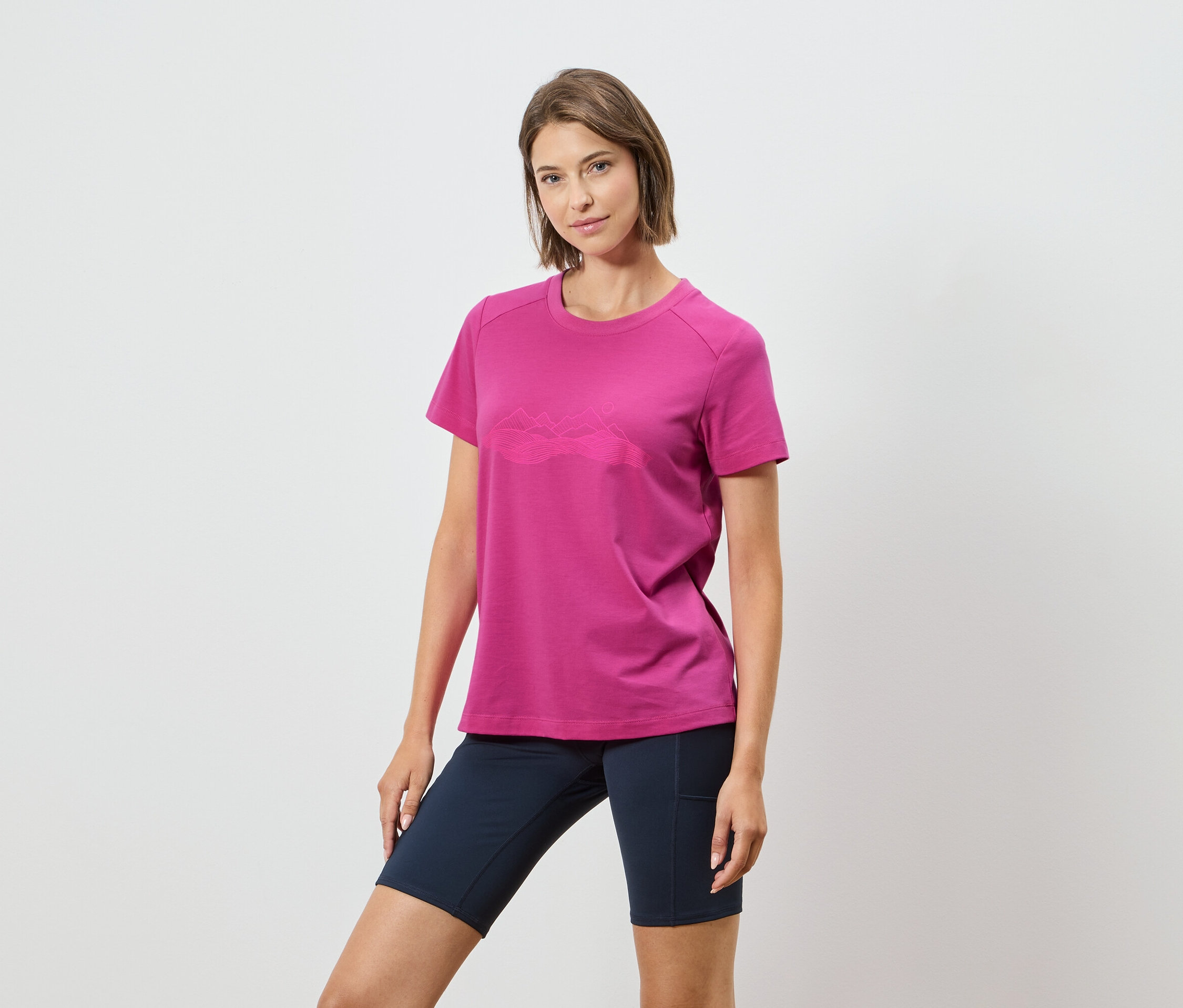Kobieta ma na sobie różową koszulkę z górskim motywem i granatowe szorty na białym tle.
