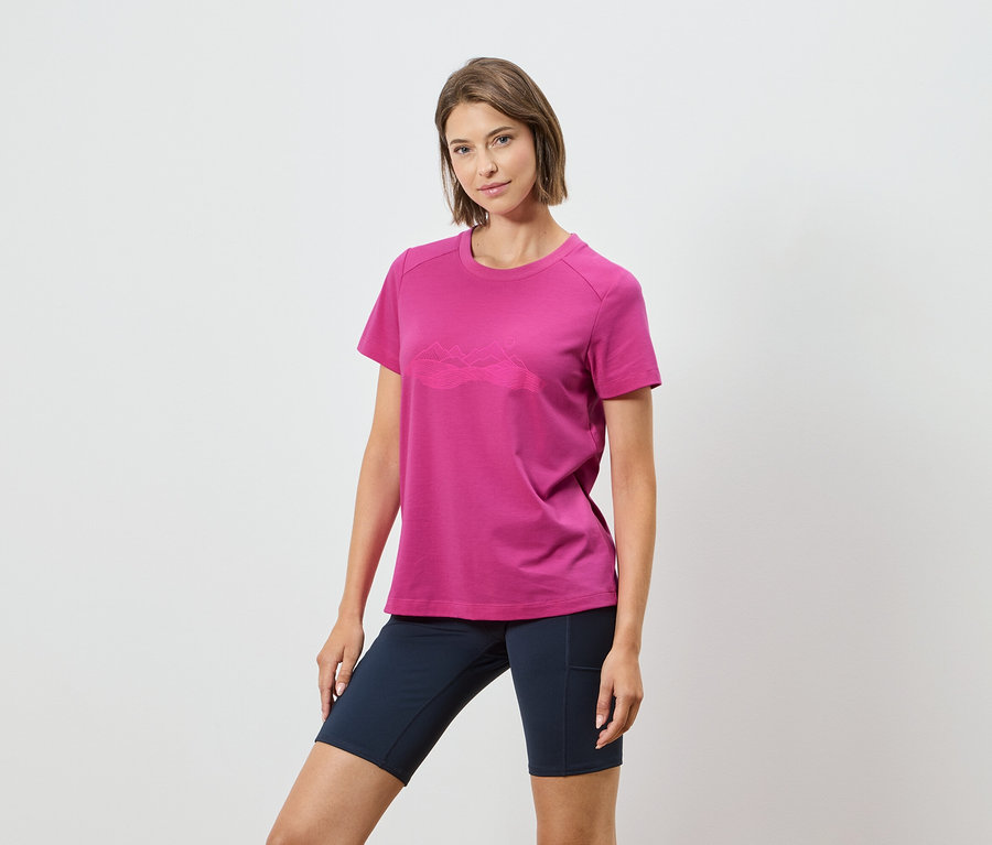 Kobieta ma na sobie różową koszulkę z górskim motywem i granatowe szorty na białym tle.