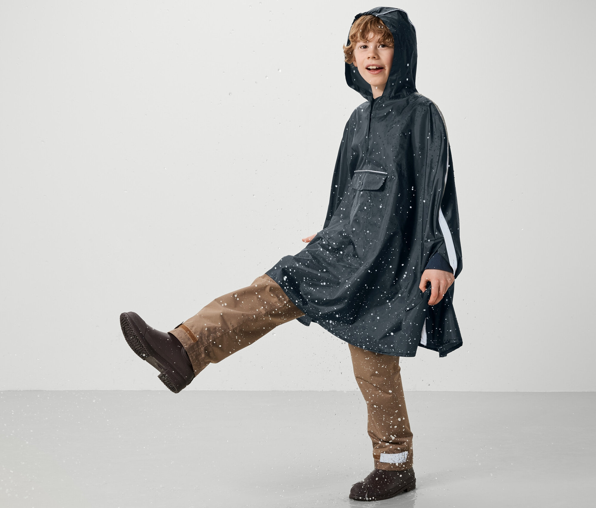 Chłopiec w niebieskim ponczo przeciwdeszczowym Kinder-Regenponcho kopie w wodzie.