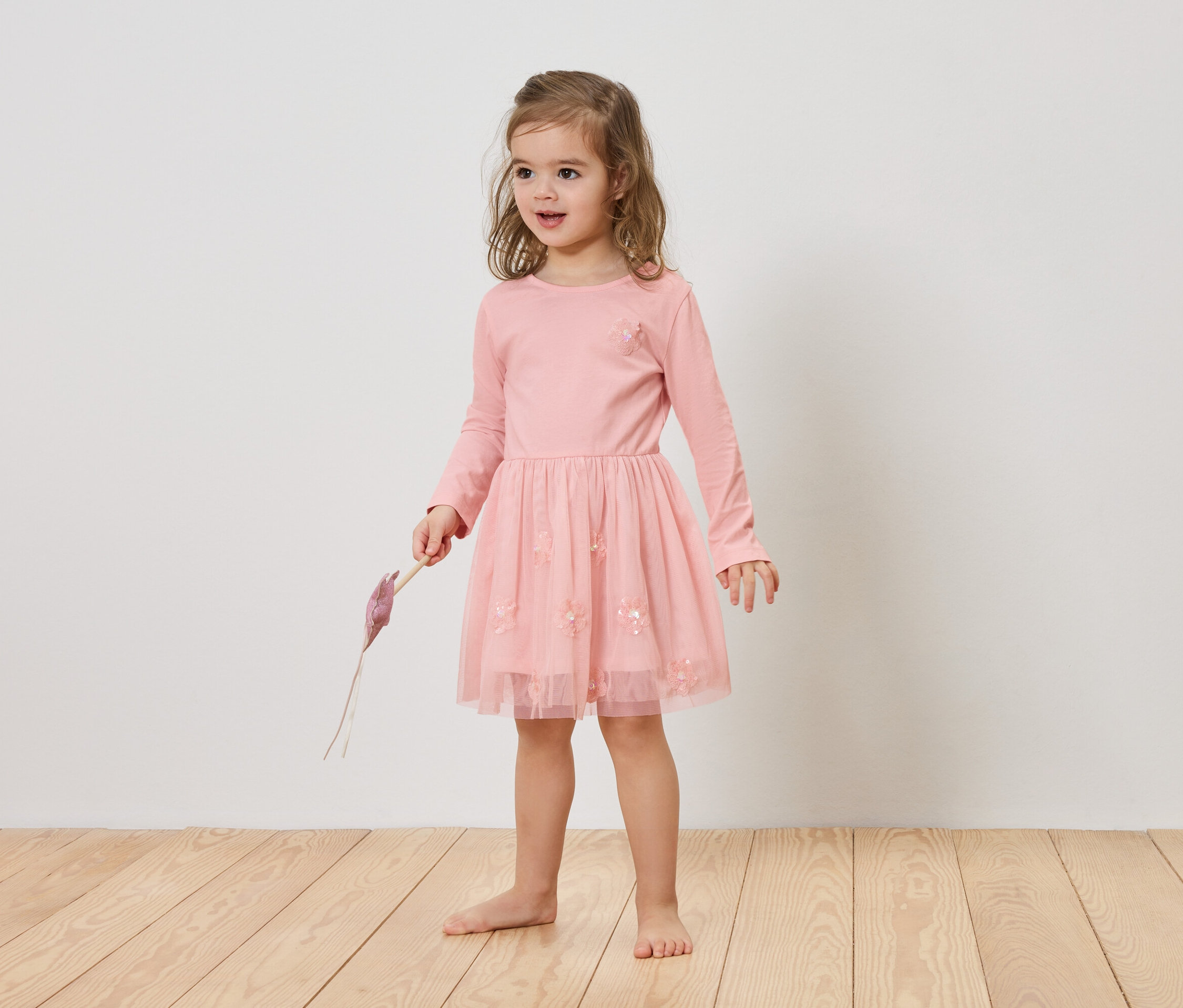 Dziewczynka z blond włosami ma na sobie różową sukienkę i trzyma w ręku fioletowy przedmiot.