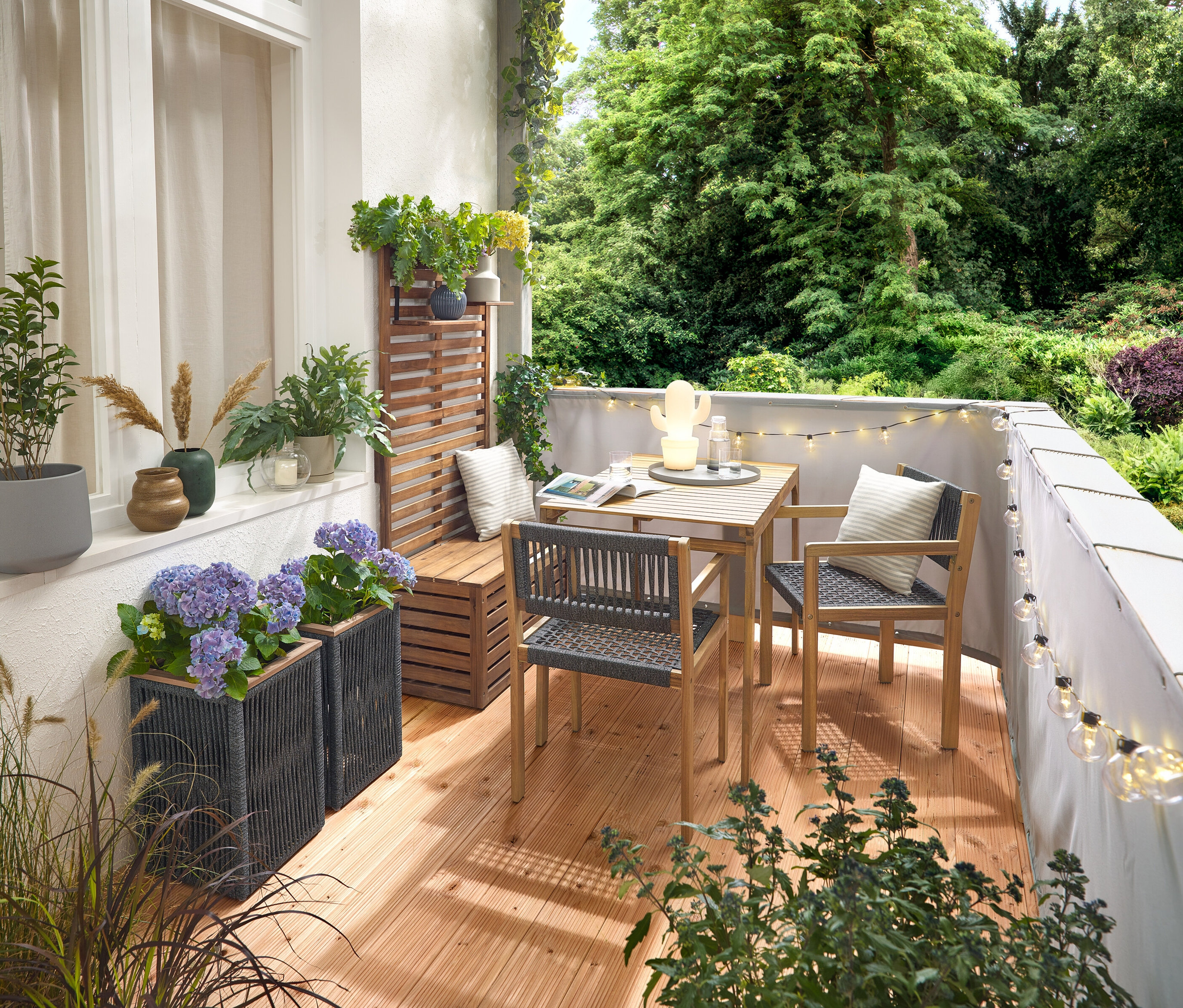 Aranżacja balkonu z zestawem balkonowym z plecionki tekstylnej, szarym parawanem balkonowym, zewnętrznym łańcuchem świetlnym LED z zasilaczem, doniczką z plecionki tekstylnej, zewnętrzną lampą stołową LED na akumulator w kształcie kaktusa i 2 poduszkami zewnętrznymi z lnu.