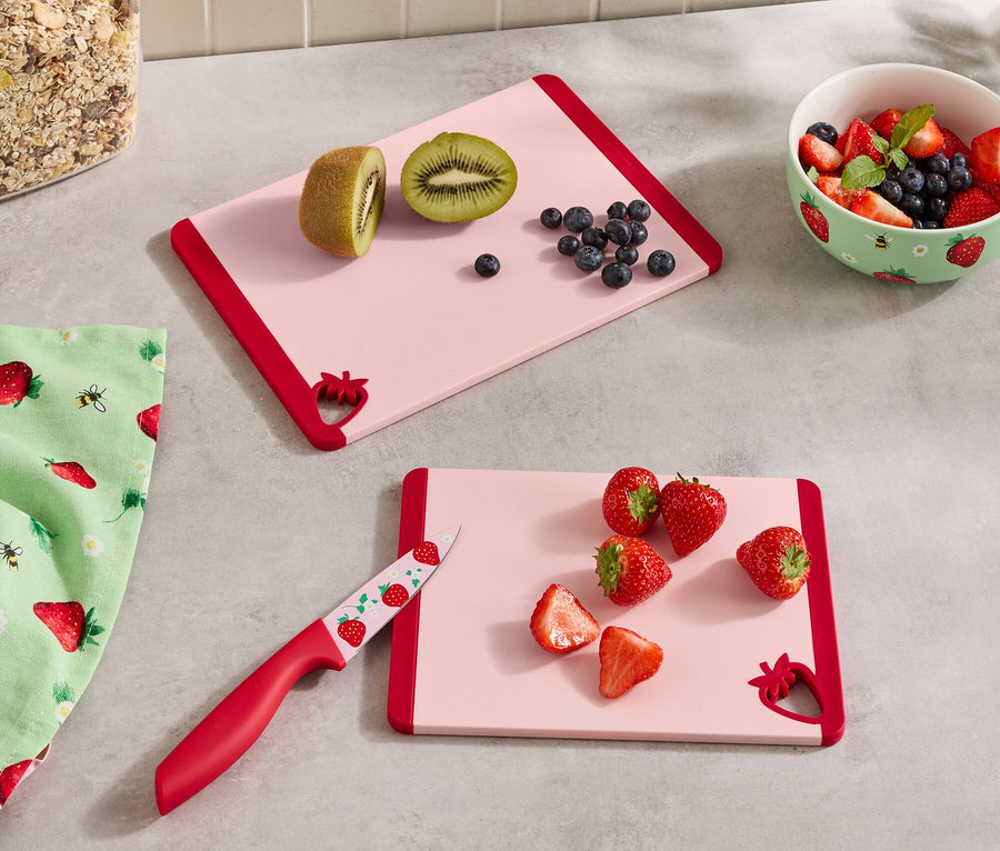 Na blacie kuchennym leżą dwie różowe deski do krojenia z czerwonymi brzegami, pojemnik z musli, miska z jagodami, ściereczka i nóż.