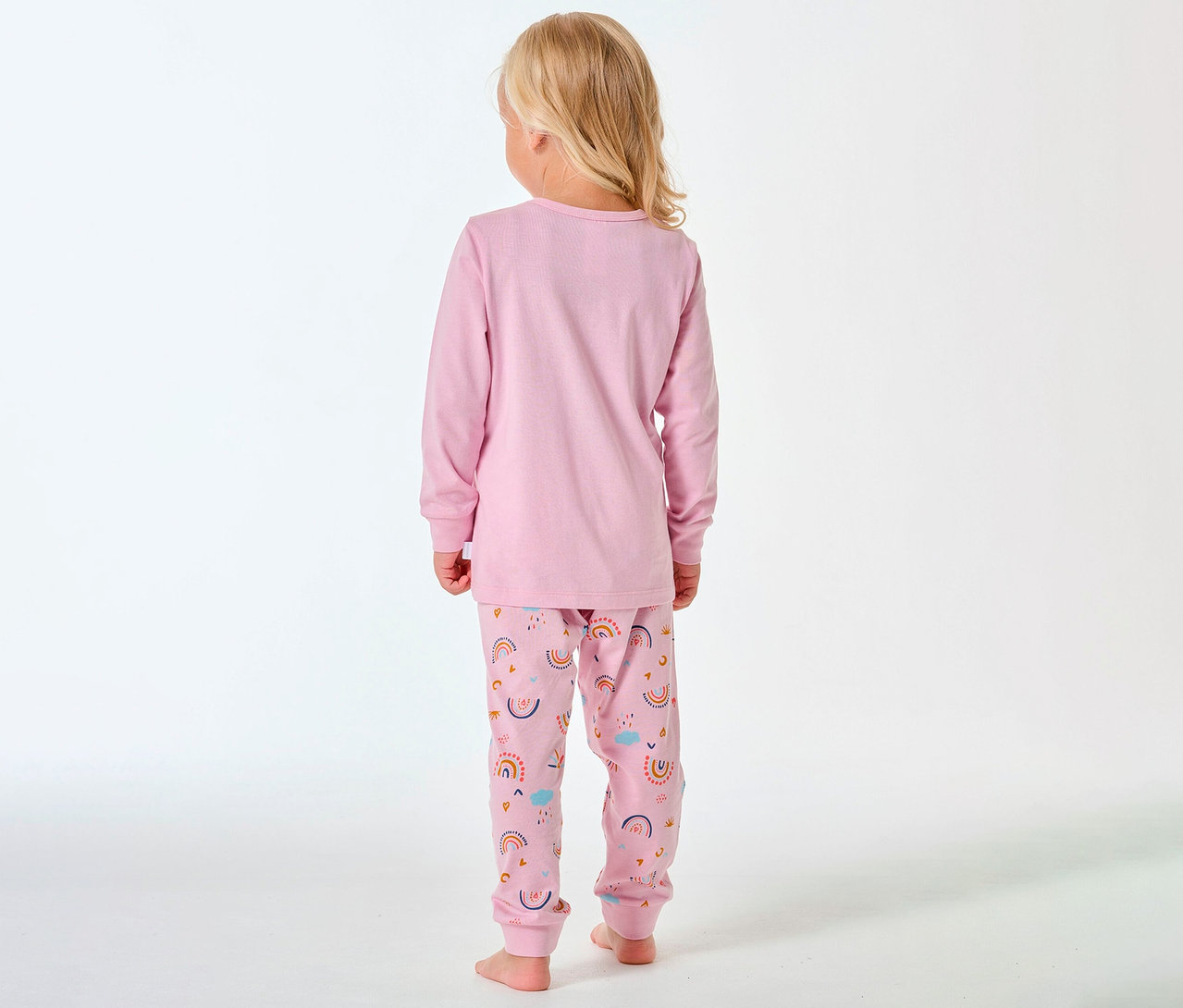 Dziewczynka z blond włosami, widziana od tyłu, ma na sobie piżamę dziecięcą SCHIESSER w tęczowy wzór.