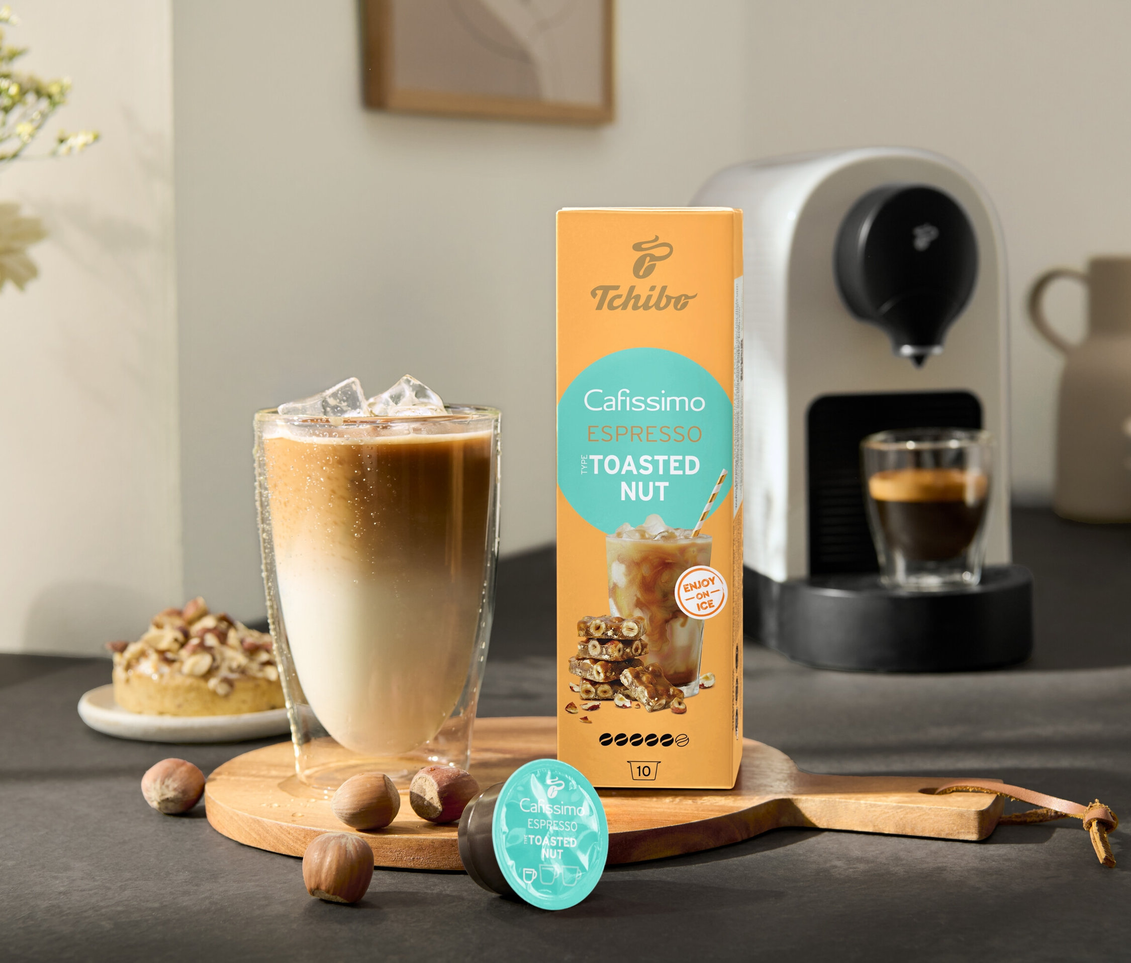 Szklanka mrożonej kawy stoi obok opakowania Cafissimo Flavoured Espresso – Toasted Nut i ekspresu do kawy Cafissimo pure+, white.