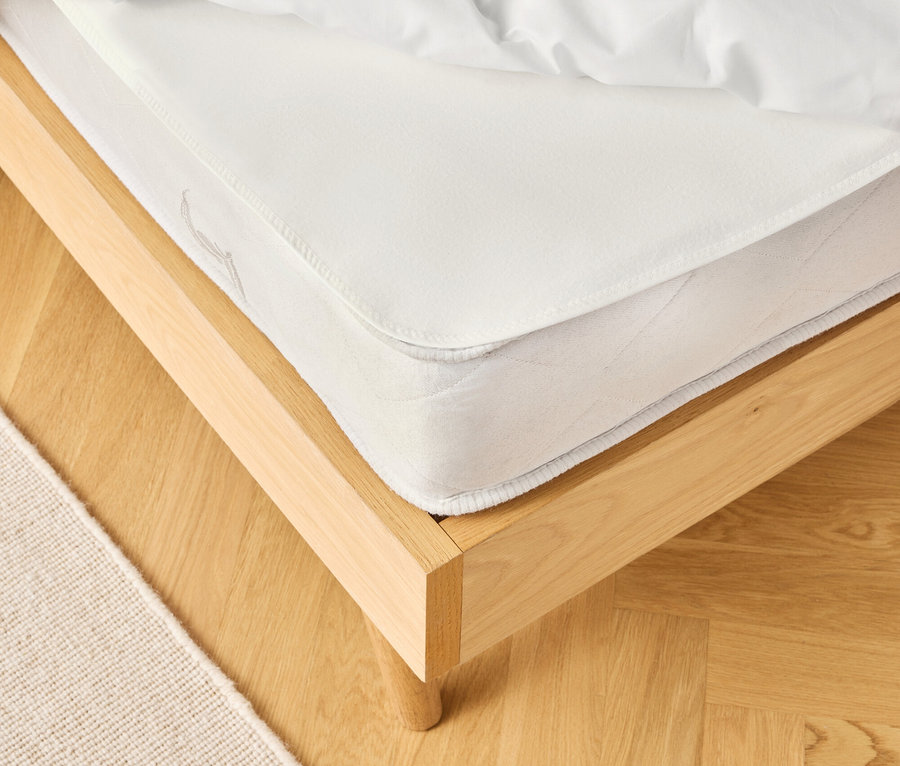 Szczegółowy widok łóżka z białym materacem i pościelą na jasnej drewnianej podłodze, obok beżowy dywan.