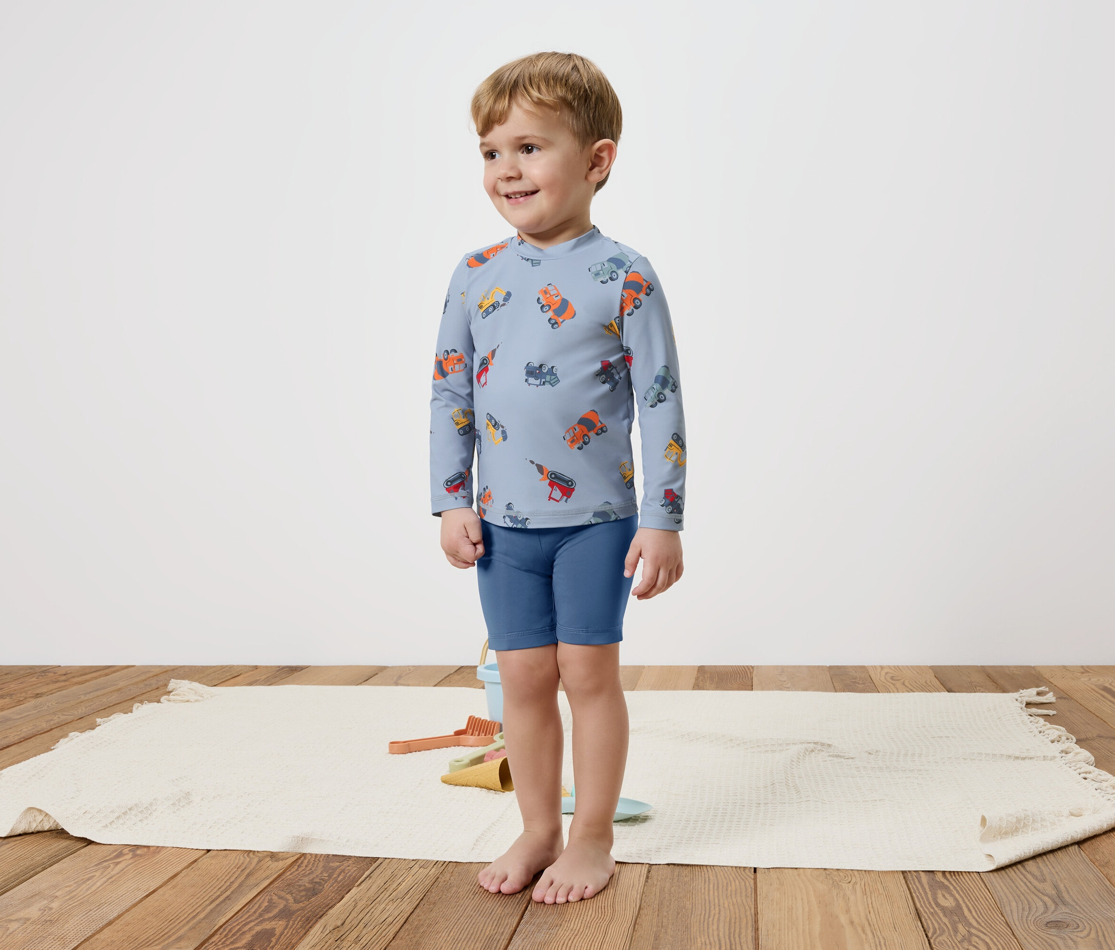 Mały chłopiec stoi boso na dywanie, ubrany w niebieską koszulkę z długim rękawem w kolorowe pojazdy i niebieskie szorty.