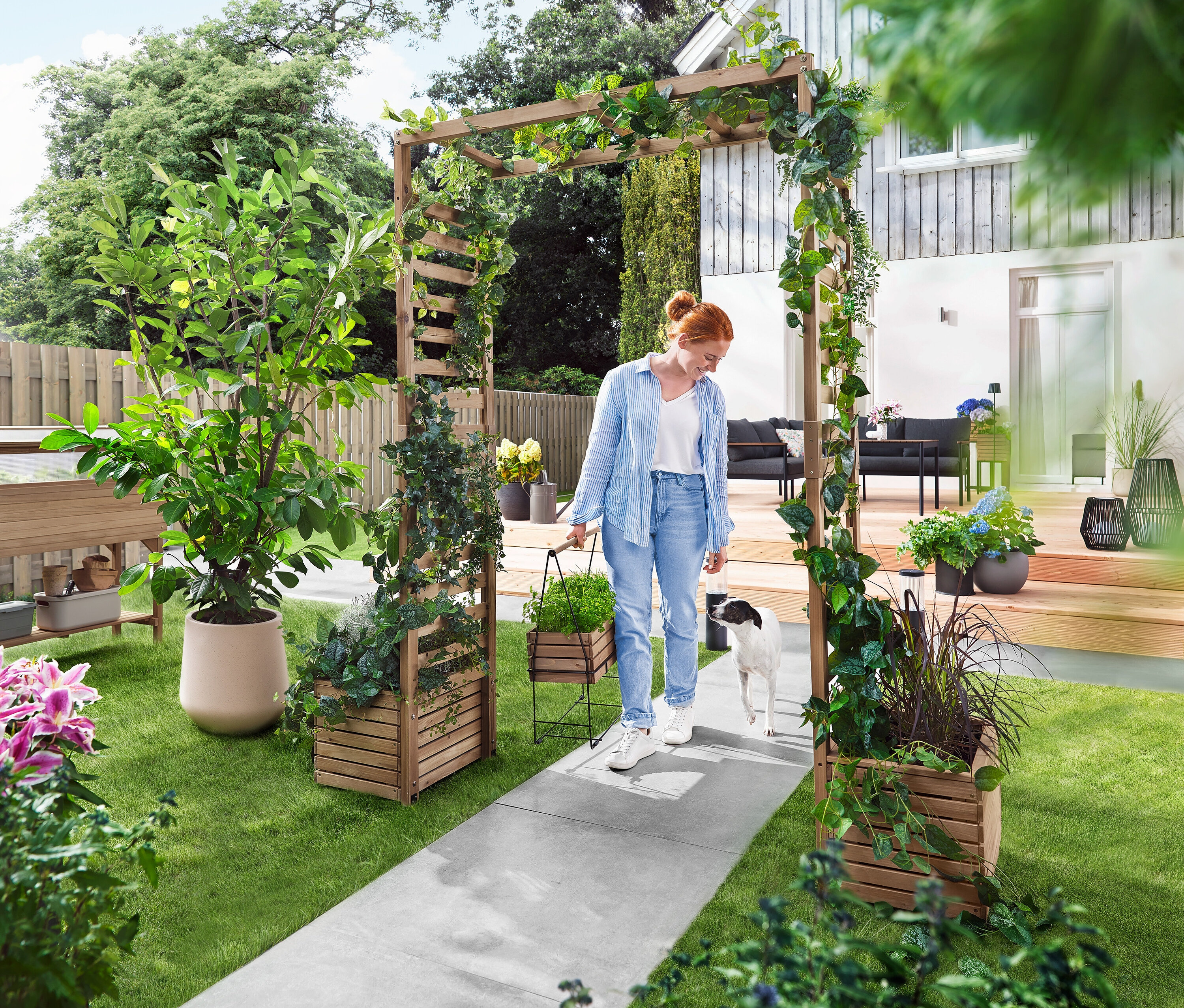 Kobieta idzie z psem przez łuk ogrodowy porośnięty roślinami. Niesie skrzynkę na rośliny.