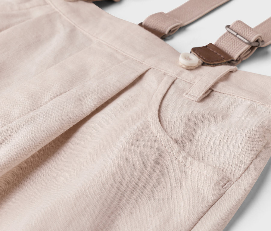 Szczegółowe ujęcie jasnobeżowych spodni na szelkach z guzikiem i kieszenią.