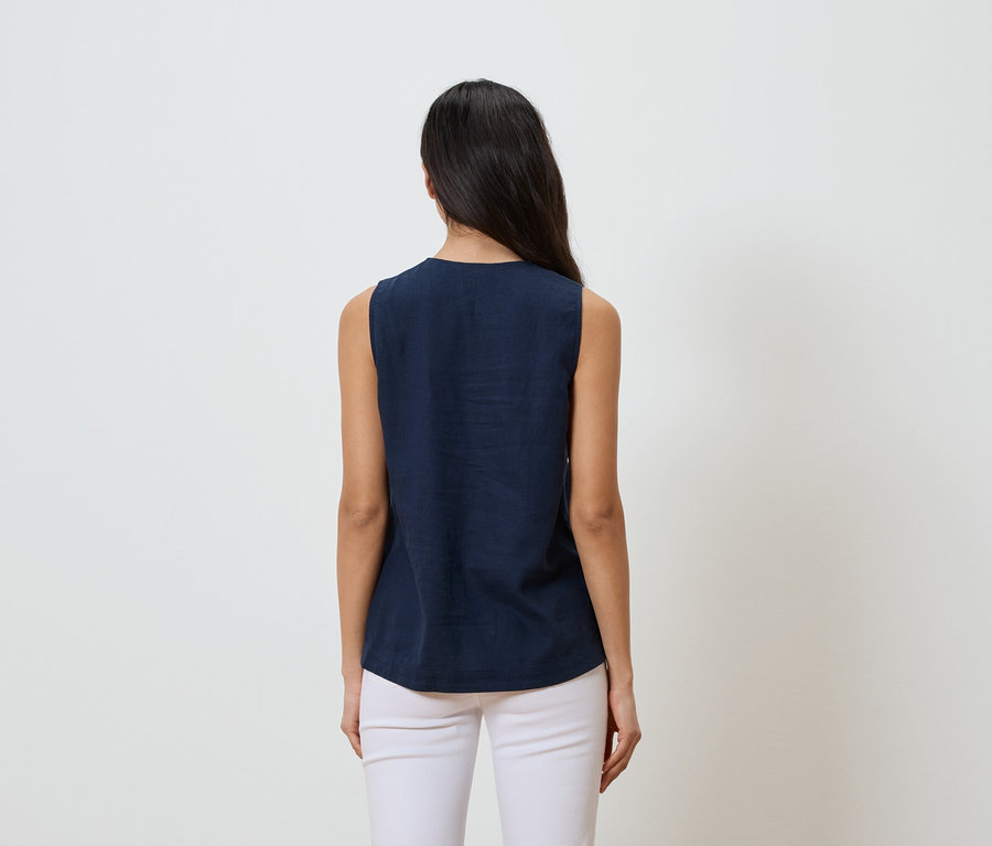 Kobieta z ciemnymi włosami, widziana od tyłu, ma na sobie granatową bluzkę bez rękawów i białe spodnie na białym tle.