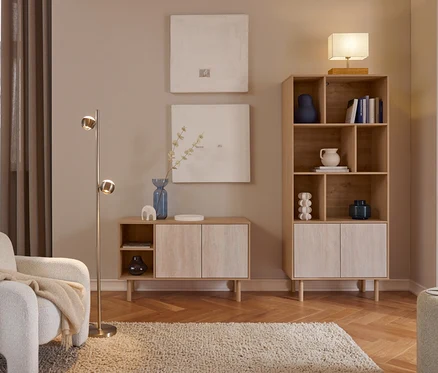 Przytulny salon z jasnymi drewnianymi meblami. Biały fotel z beżowym kocem stoi obok mosiężnej lampy podłogowej. Dwa abstrakcyjne obrazy wiszą nad niską szafką z niebieskim wazonem. Wysoki regał na książki z dekoracjami i książkami jest zwieńczony lampą stołową. Kudłaty dywan leży na parkiecie w jodełkę.