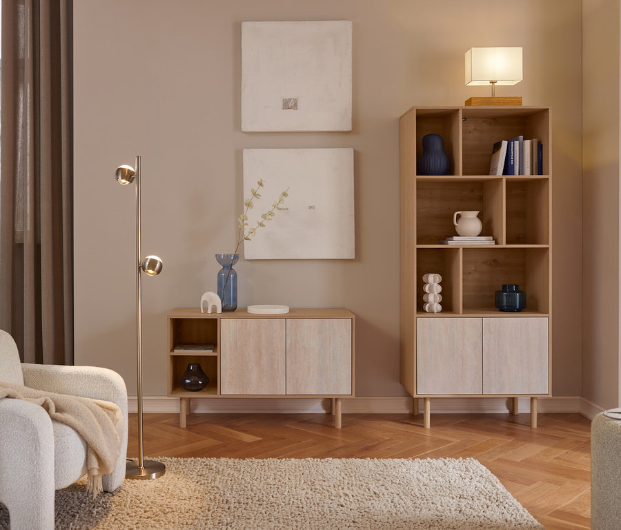 Przytulny salon z jasnymi drewnianymi meblami. Biały fotel z beżowym kocem stoi obok mosiężnej lampy podłogowej. Dwa abstrakcyjne obrazy wiszą nad niską szafką z niebieskim wazonem. Wysoki regał na książki z dekoracjami i książkami jest zwieńczony lampą stołową. Kudłaty dywan leży na parkiecie w jodełkę.