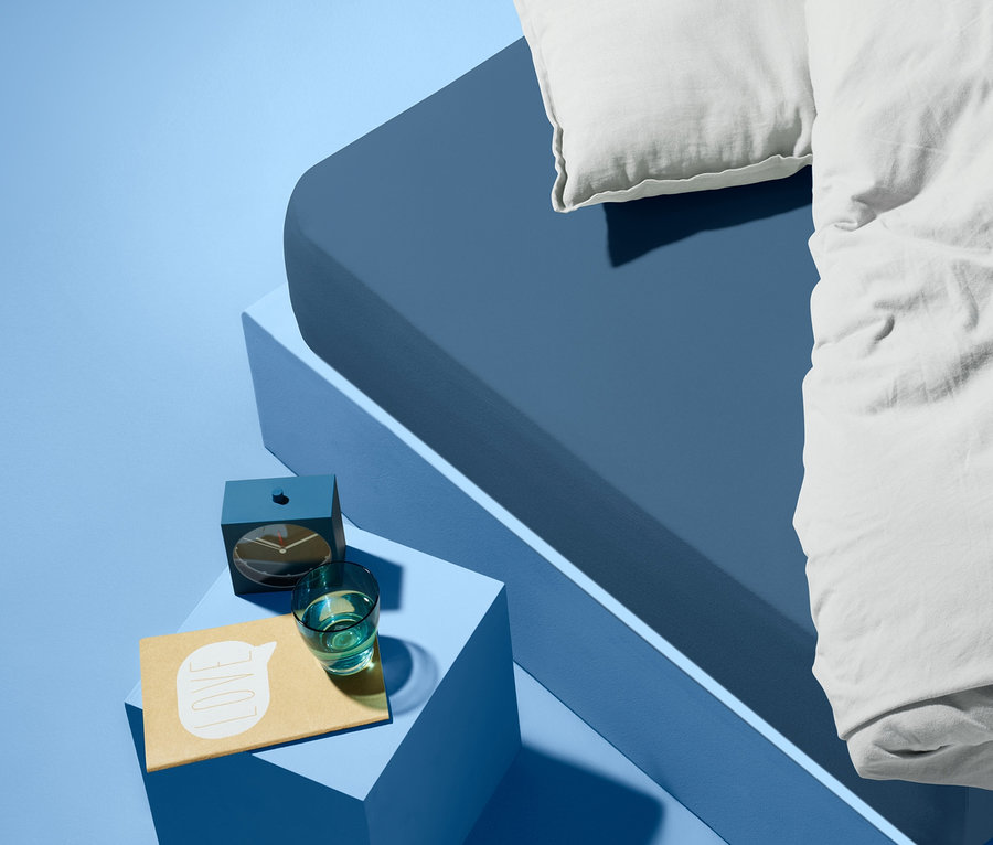 Niebieskie łóżko z białą poduszką i kołdrą. Na niebieskim stoliku nocnym obok łóżka stoi budzik, szklanka wody i notatnik.