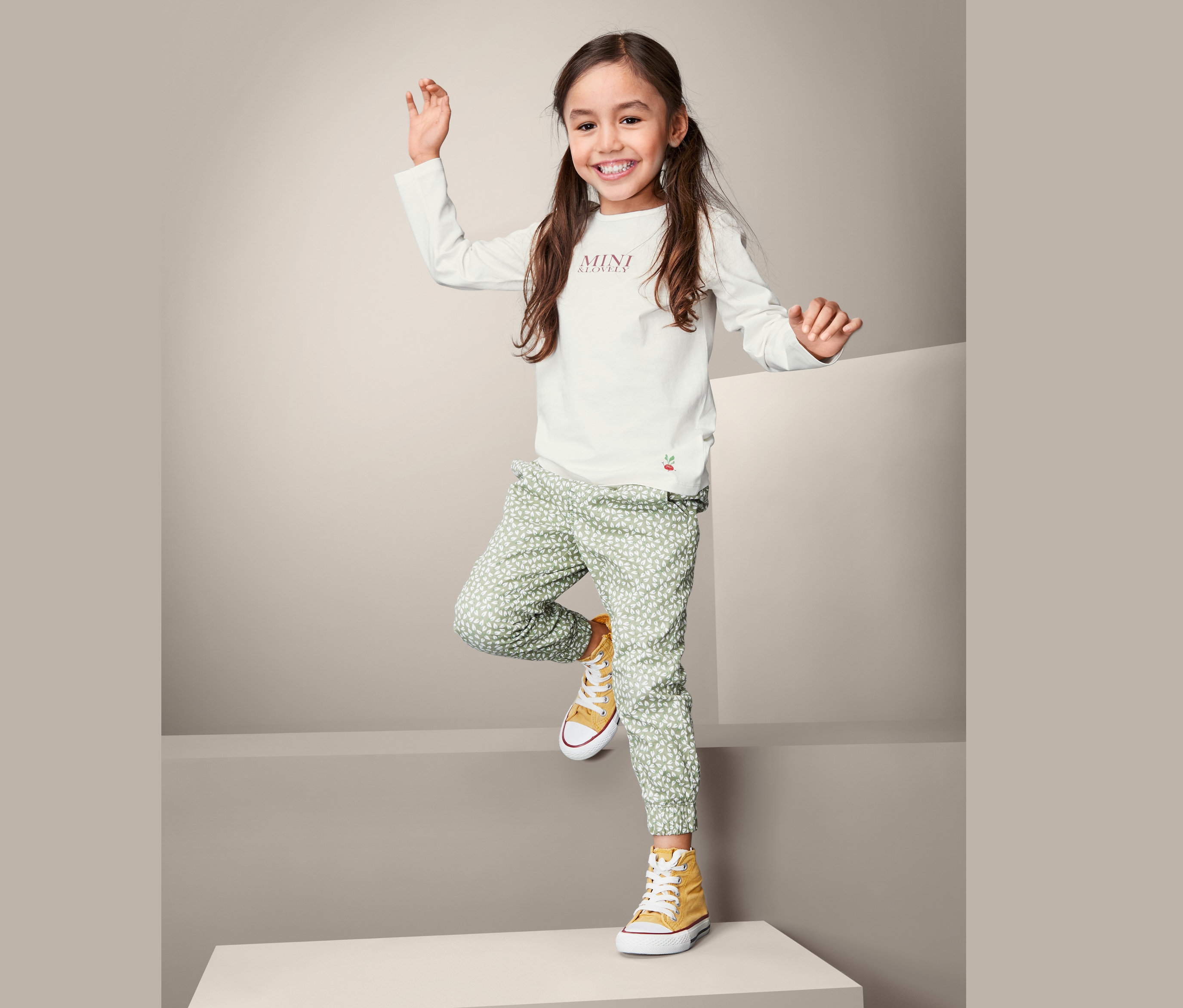 Uśmiechnięta dziewczyna pozuje w koszulce z długim rękawem, zielonych wzorzystych spodniach i żółtych trampkach.