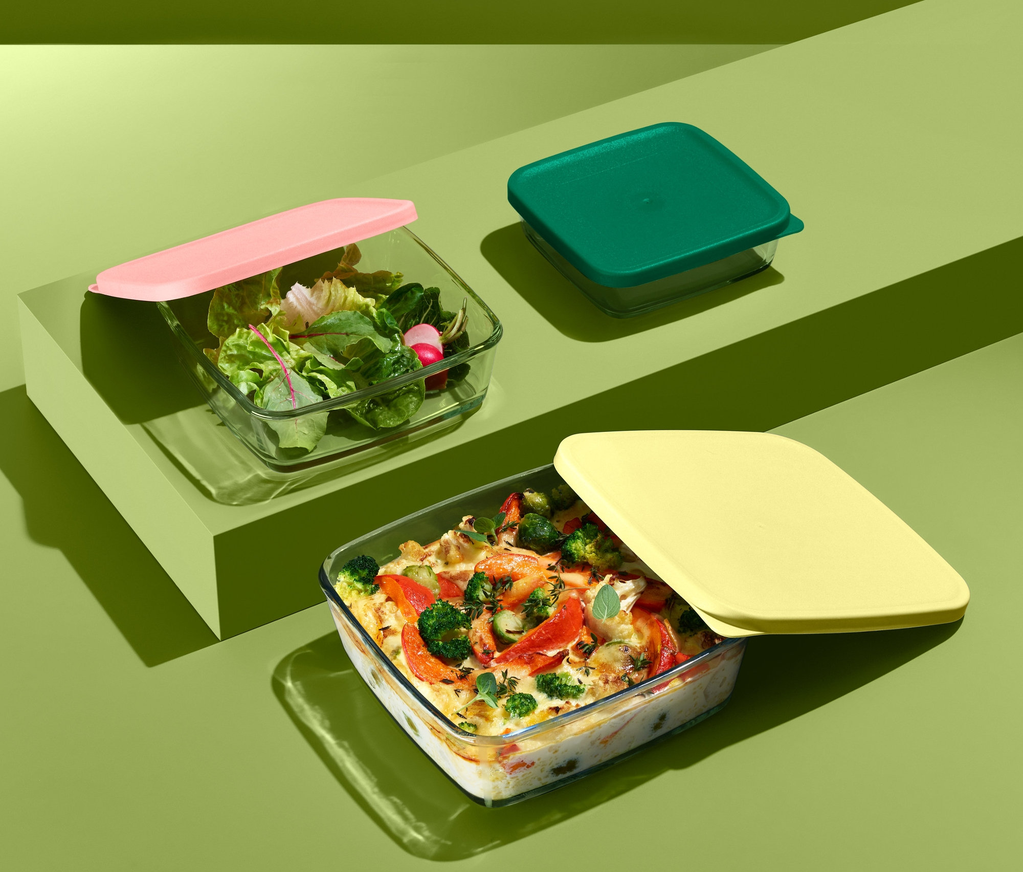 Trzy szklane formy do zapiekania z pokrywką. Jedna z różową pokrywką i sałatką, jedna z zieloną pokrywką, jedna z żółtą pokrywką i jedzeniem.