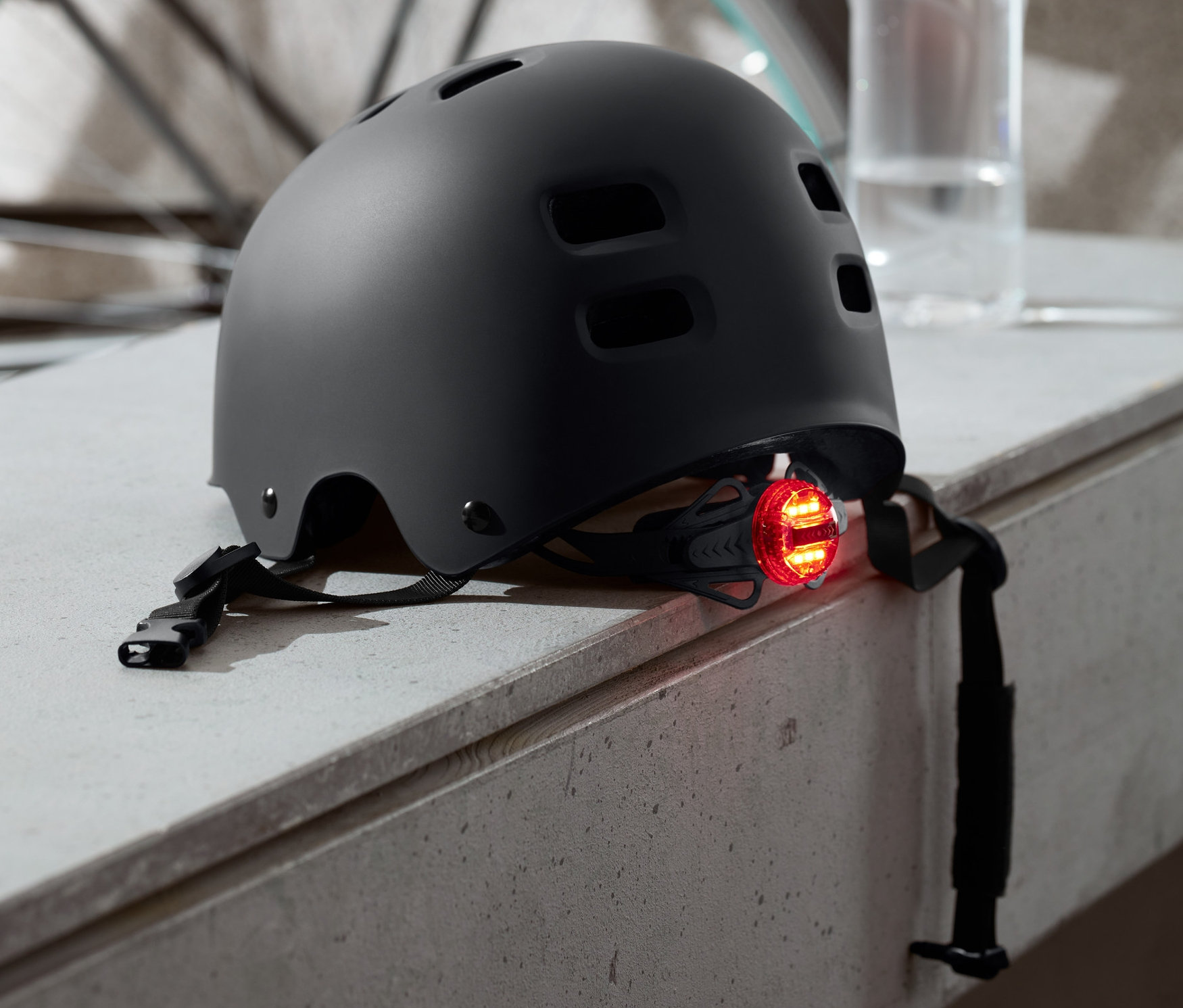 Czarny kask rowerowy z tylnym światłem LED leży na murku, w tle rower i butelka z wodą.