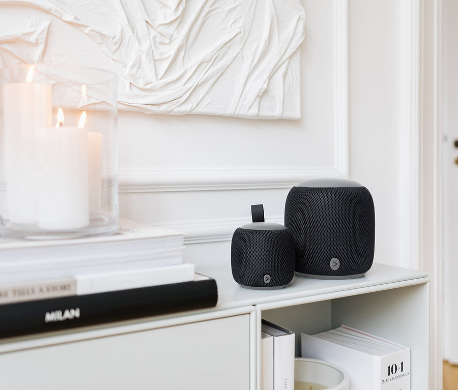 Dwa czarne głośniki Fabric Bluetooth® w różnych rozmiarach stoją na białej półce obok świec i książek.