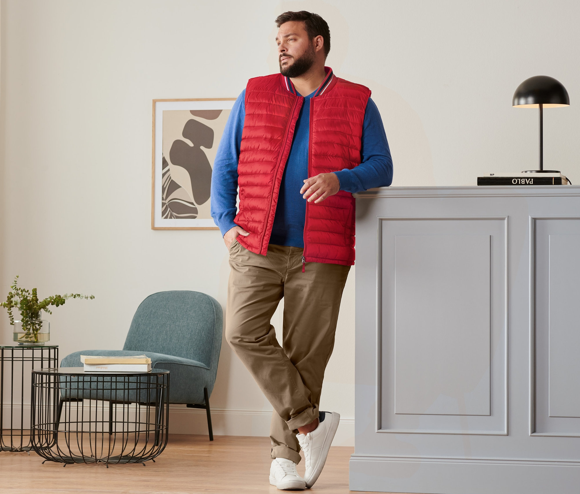 Mężczyzna opiera się o komodę, ubrany w czerwoną kamizelkę pikowaną, niebieski sweter lniany, karmelowe spodnie chino i buty typu sneaker.