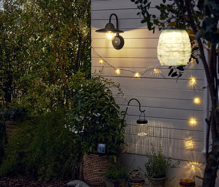 Na ścianie wisi akumulatorowy kinkiet LED z dodatkowym akumulatorem. Obok niego znajduje się solarny łańcuch świetlny. Na pierwszym planie znajduje się lampion solarny i lampa solarna na stelażu.