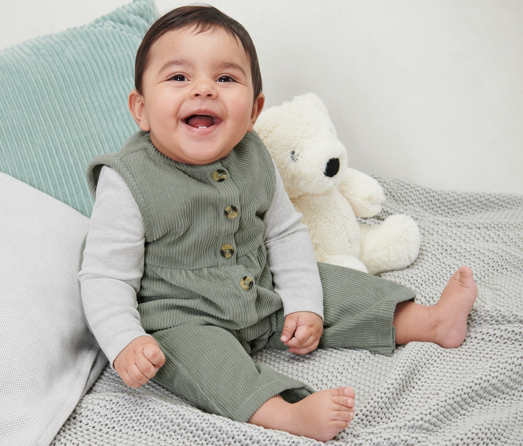 Uśmiechnięte dziecko siedzi na szarym kocu obok pluszowego misia, ubrane w dziecięce ogrodniczki sztruksowe.