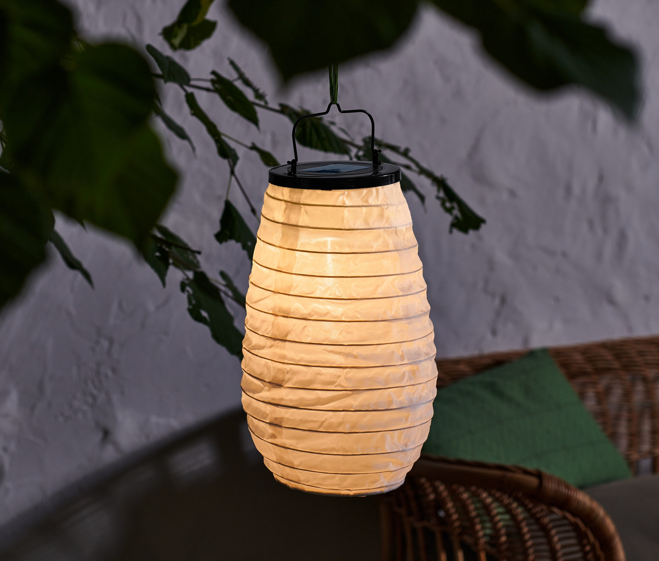 Wiszący lampion solarny z czarnym uchwytem, umieszczony na zewnątrz wśród gałęzi drzewa.