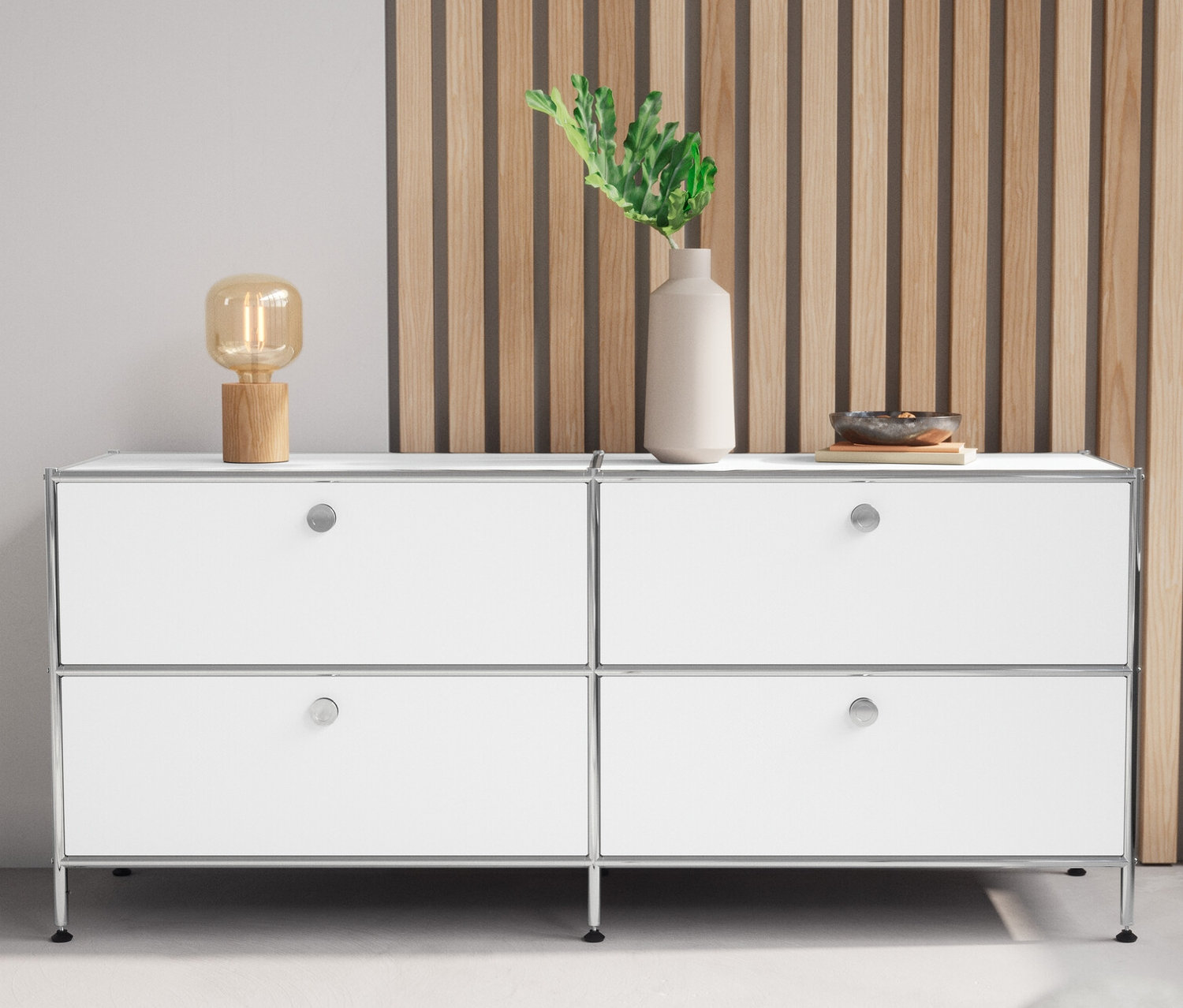 Biała niska szafka metalowa »CN3« z 4 szufladami i drewniana lampa stołowa stoją obok siebie.