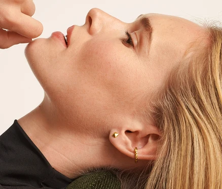 Kobieta leży na zielonym krześle i dotyka ust. Ma na sobie srebrne kolczyki 925 piramida i srebrne kolczyki 925 ear cuff piramida.