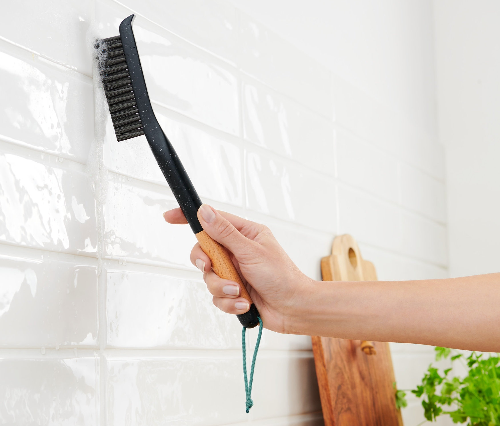 Kobieta czyści białe płytki długą szczotką do fug.