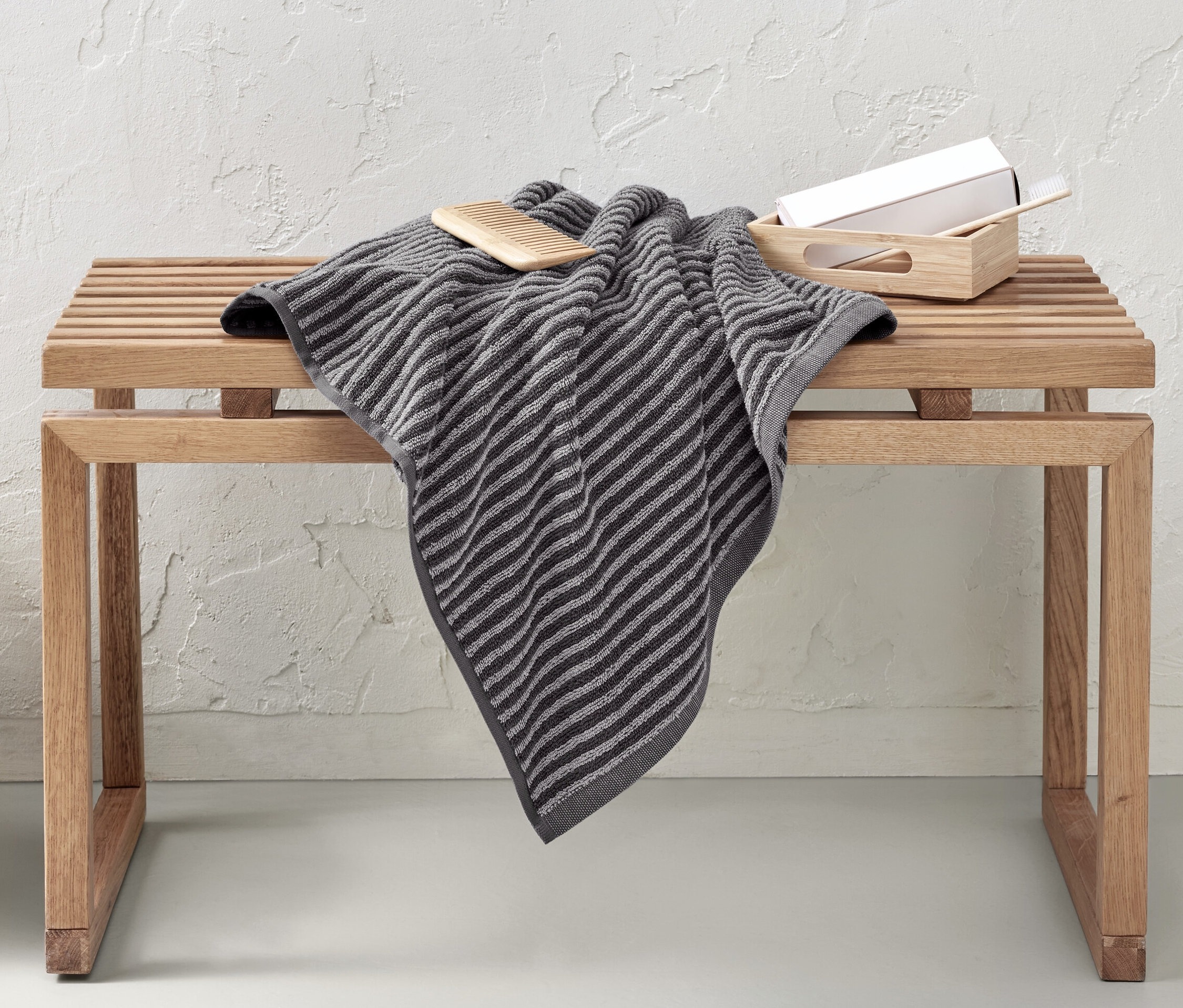 Na drewnianej ławce leży wysokiej jakości szary ręcznik kąpielowy. Obok ławki stoi wiklinowy kosz.
