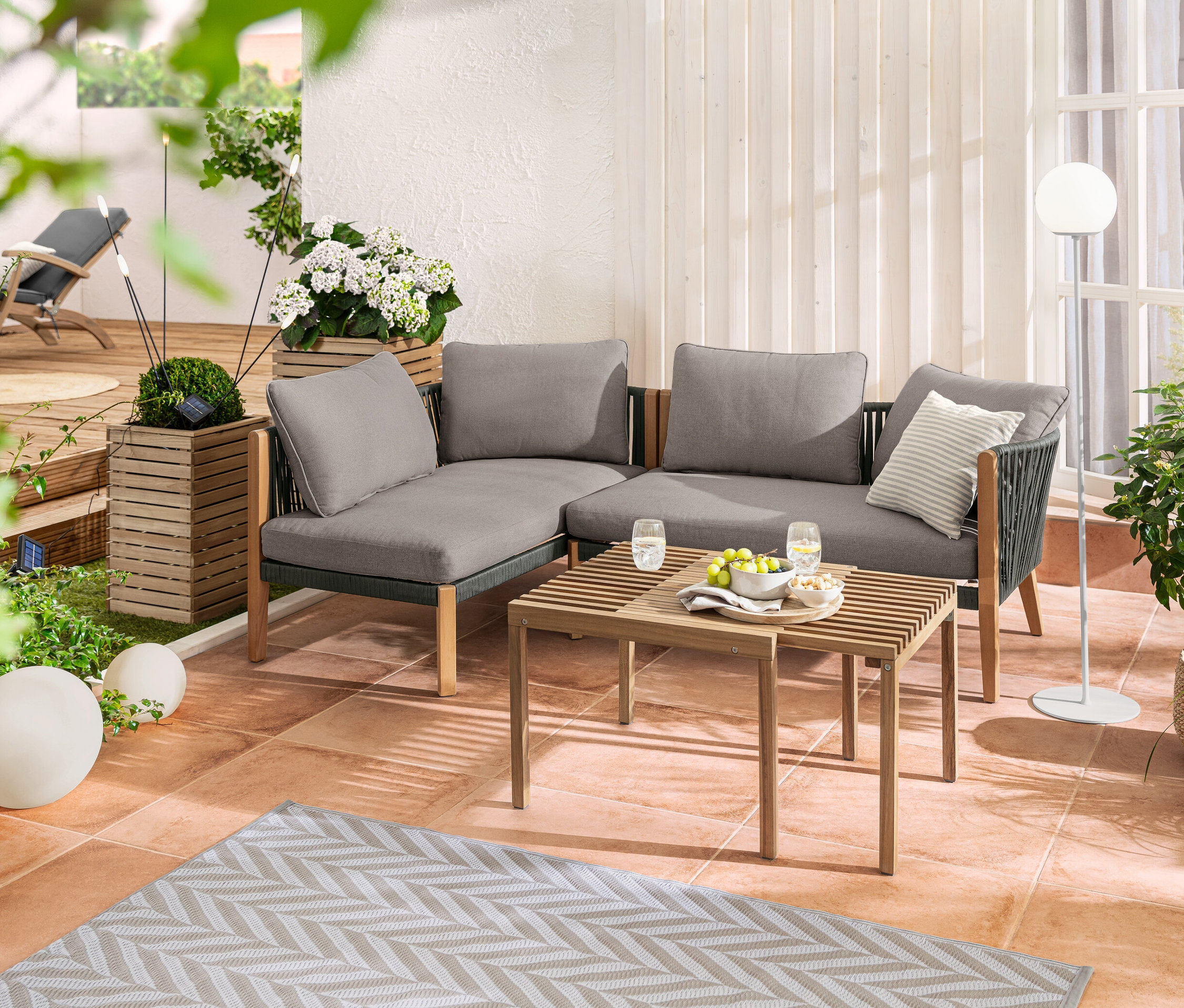 Narożnik wypoczynkowy z plecionką tekstylną na tarasie. Uzupełniają go lampy solarne w kształcie kuli (20 i 30 cm), lampa LED i zestaw donic „Tinus”.