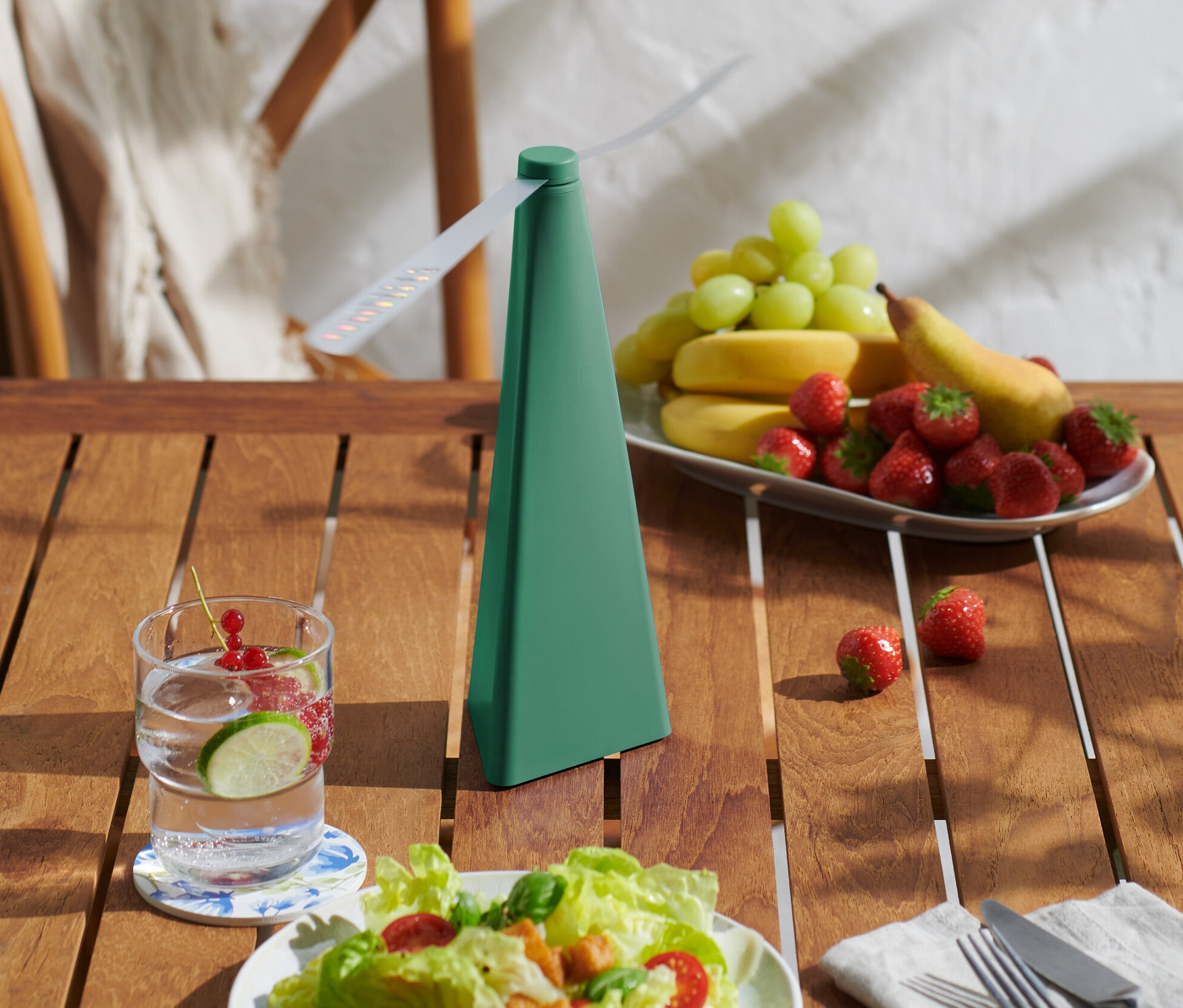 Na stole stoi odstraszacz much obok szklanki wody z owocami i talerza owoców.