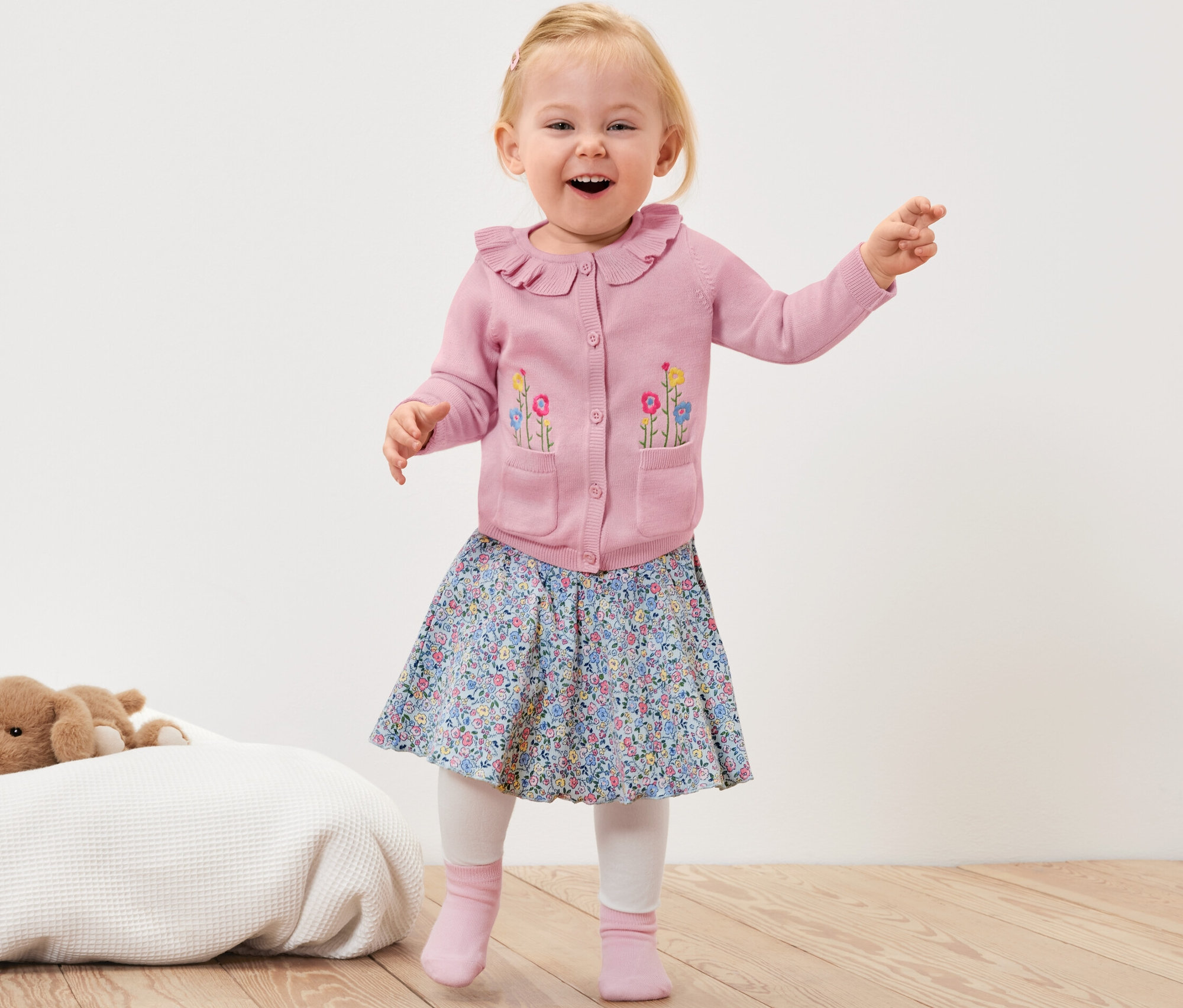 Uśmiechnięte dziecko macha rękami, ma na sobie różowy rozpinany sweter w kwiaty, kwiecistą spódnicę i różowe skarpetki.