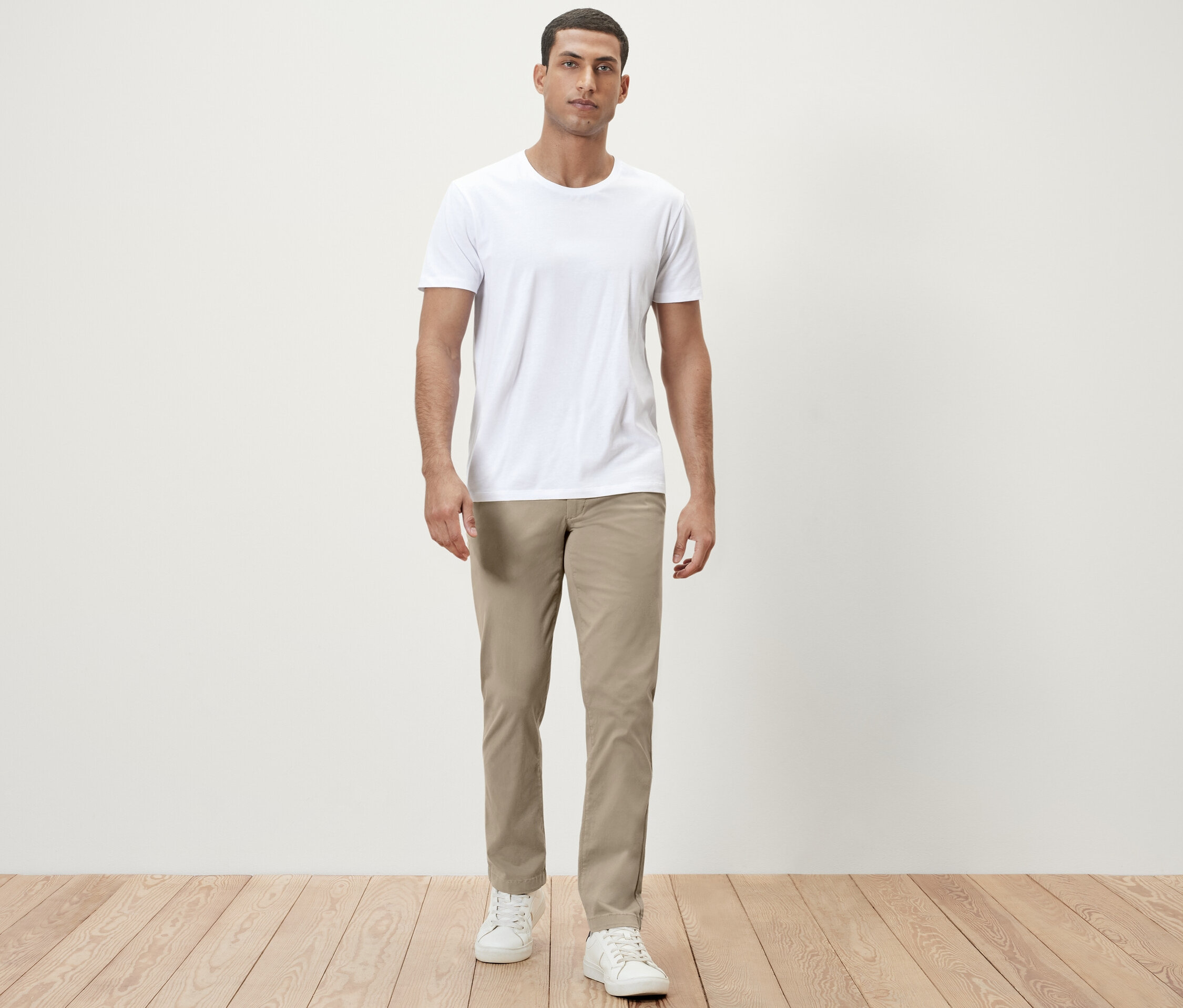 Mężczyzna stoi w białej koszulce i beżowych spodniach typu chinosy.