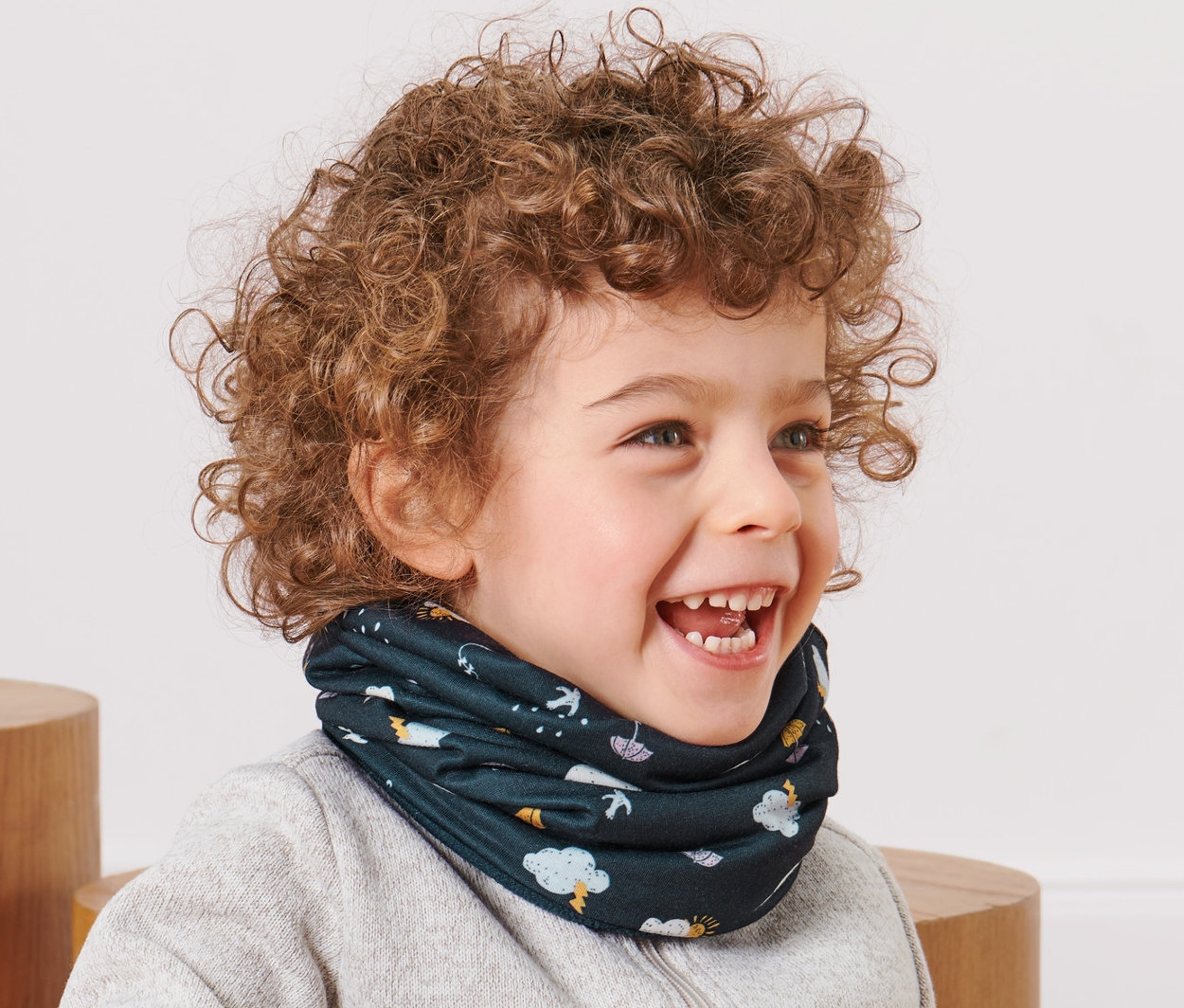 Uśmiechnięty mały chłopiec z kręconymi włosami siedzi i ma na sobie wielofunkcyjną chustkę.