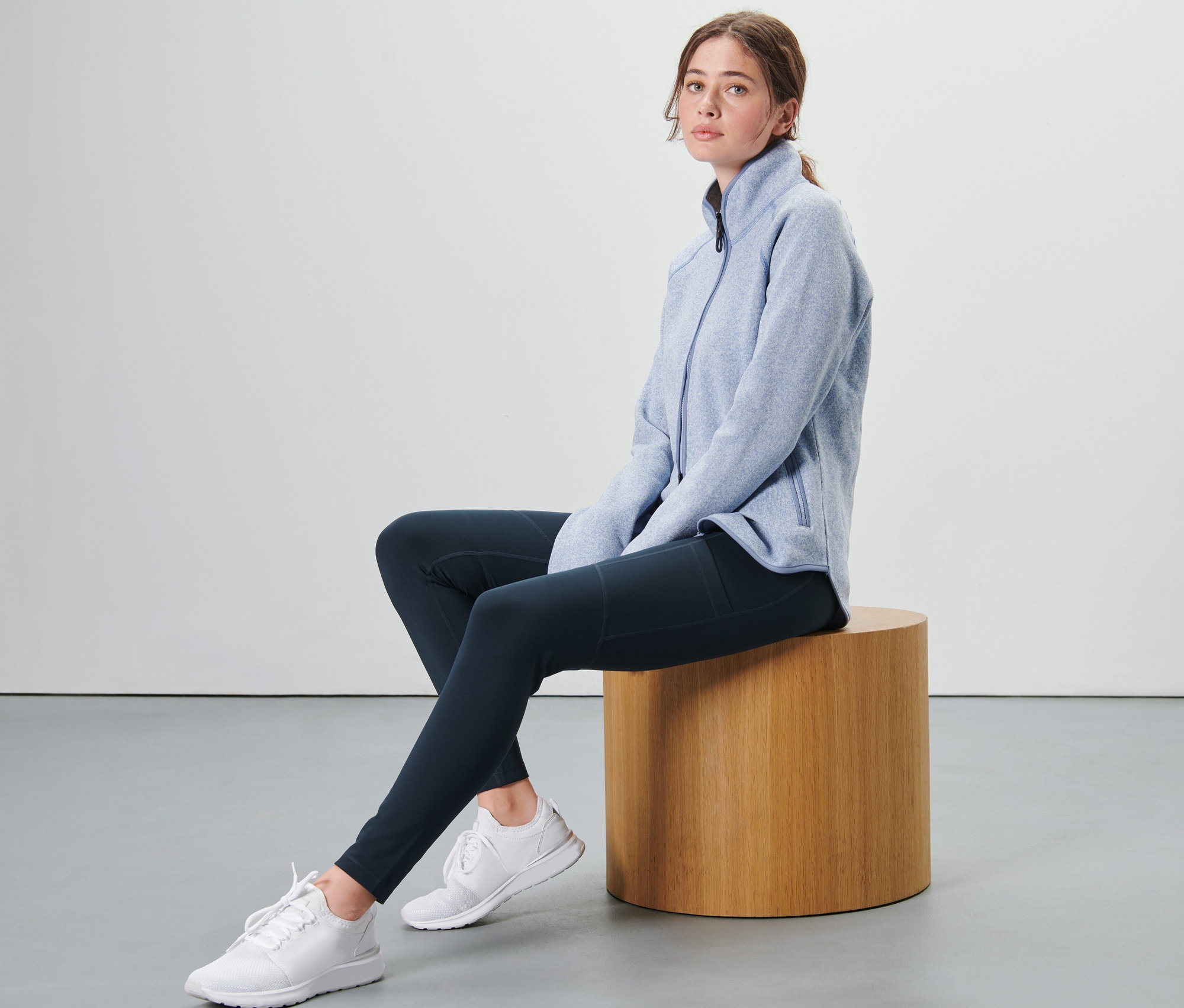 Kobieta siedzi na drewnianym stołku, ubrana w niebieską bluzę polarową i legginsy trekkingowe.