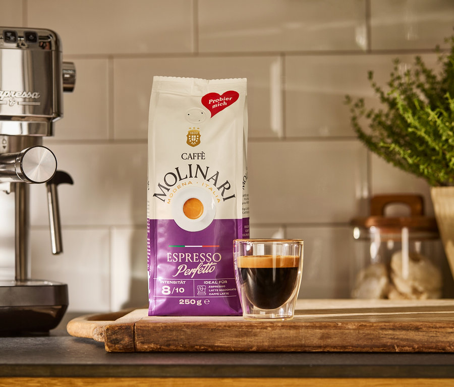 Srebrny ekspres kolbowy do kawy »Lapressa«, paczka Caffè Molinari i filiżanka espresso na drewnianej desce.