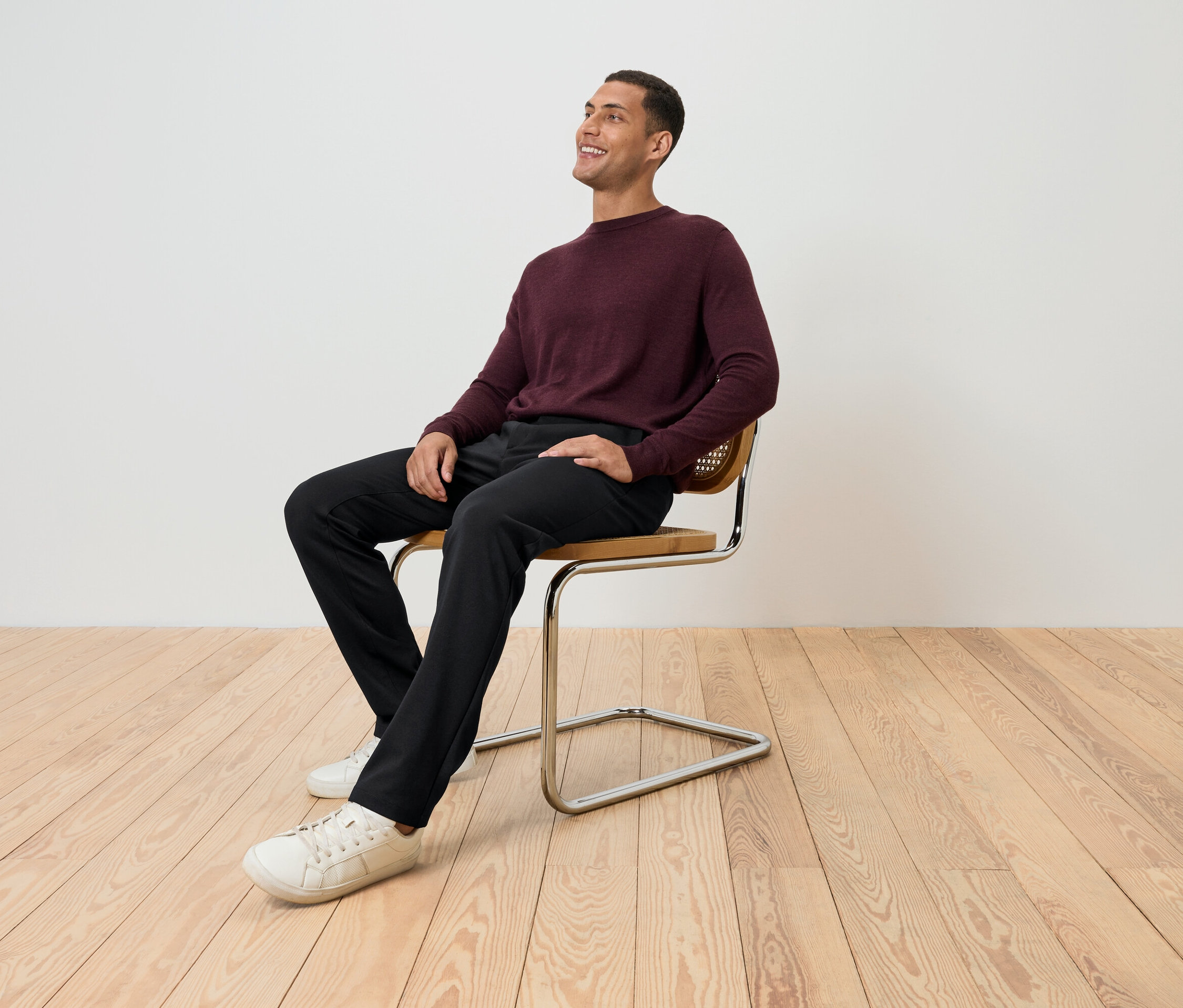 Mężczyzna siedzi na krześle w bordowym swetrze, czarnych spodniach i białych butach, patrząc w górę.