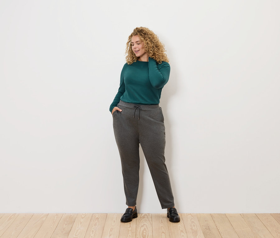 Kobieta z kręconymi włosami pozuje na jasnym tle. Ma na sobie zieloną bluzkę z długim rękawem i szare spodnie dresowe.