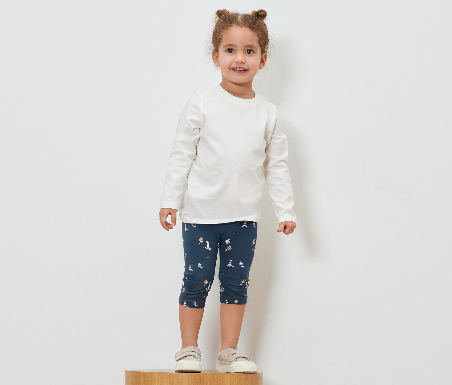 Dziewczynka stoi na drewnianym cokole, ubrana w marynistyczne legginsy 3/4.