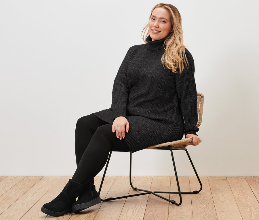 Kobieta siedzi na krześle i ma na sobie długi, czarny sweter z warkoczowym wzorem.