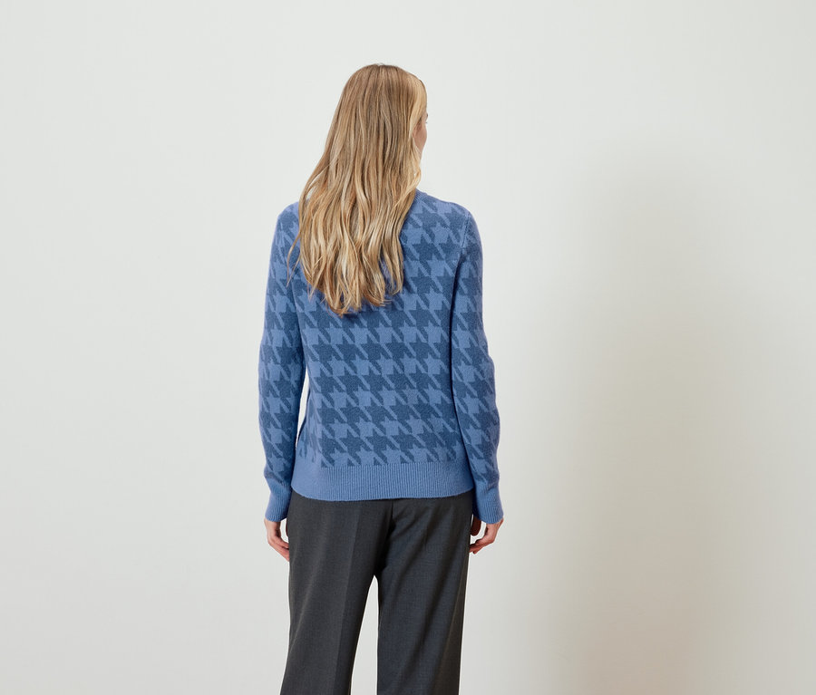 Kobieta z blond włosami, widziana z tyłu, ma na sobie niebieski sweter w kurzą stopkę i szare spodnie.